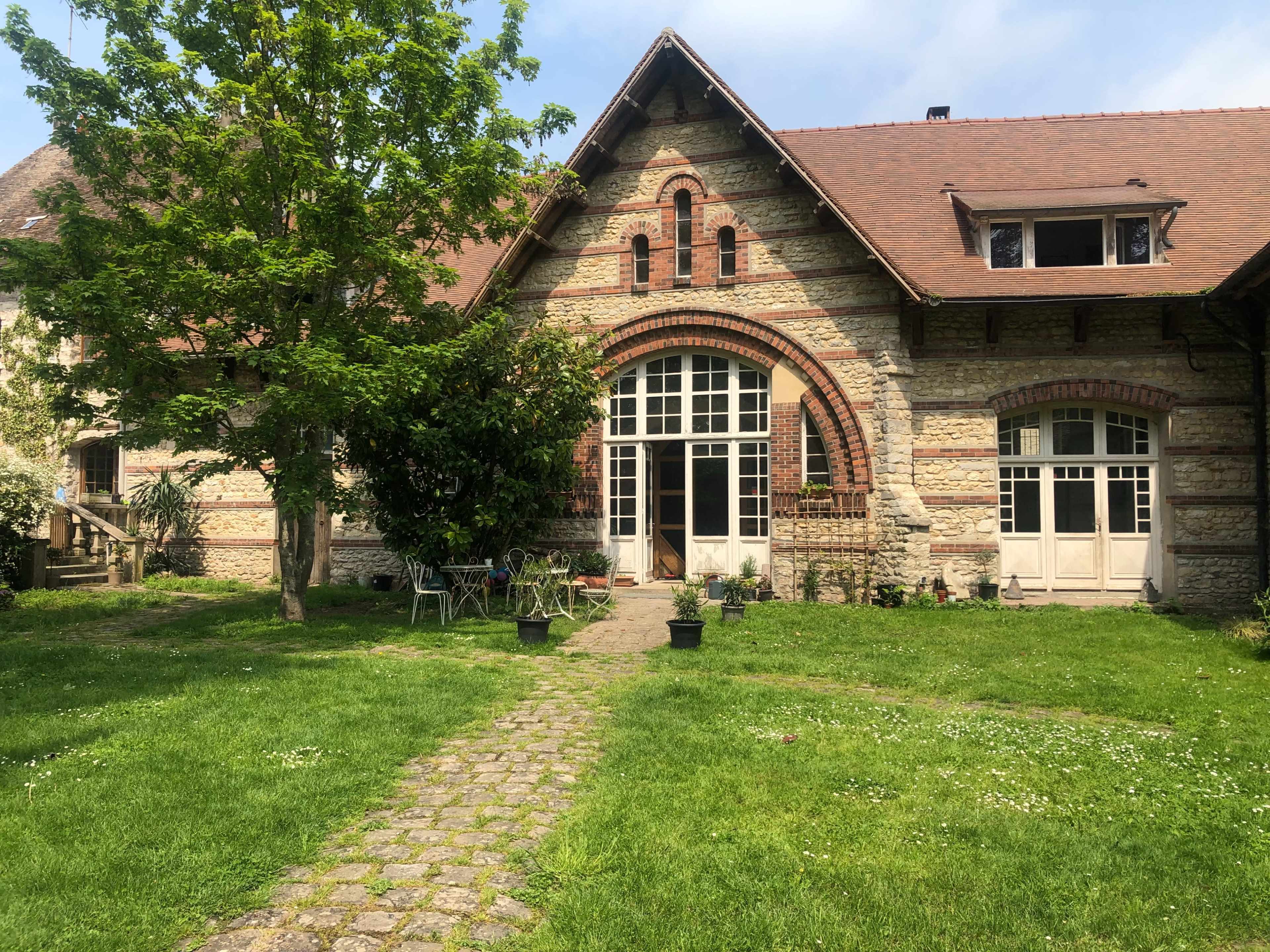 A stone and brick house with large windows and a prominent archway is surrounded by a grassy area and several potted plants.
