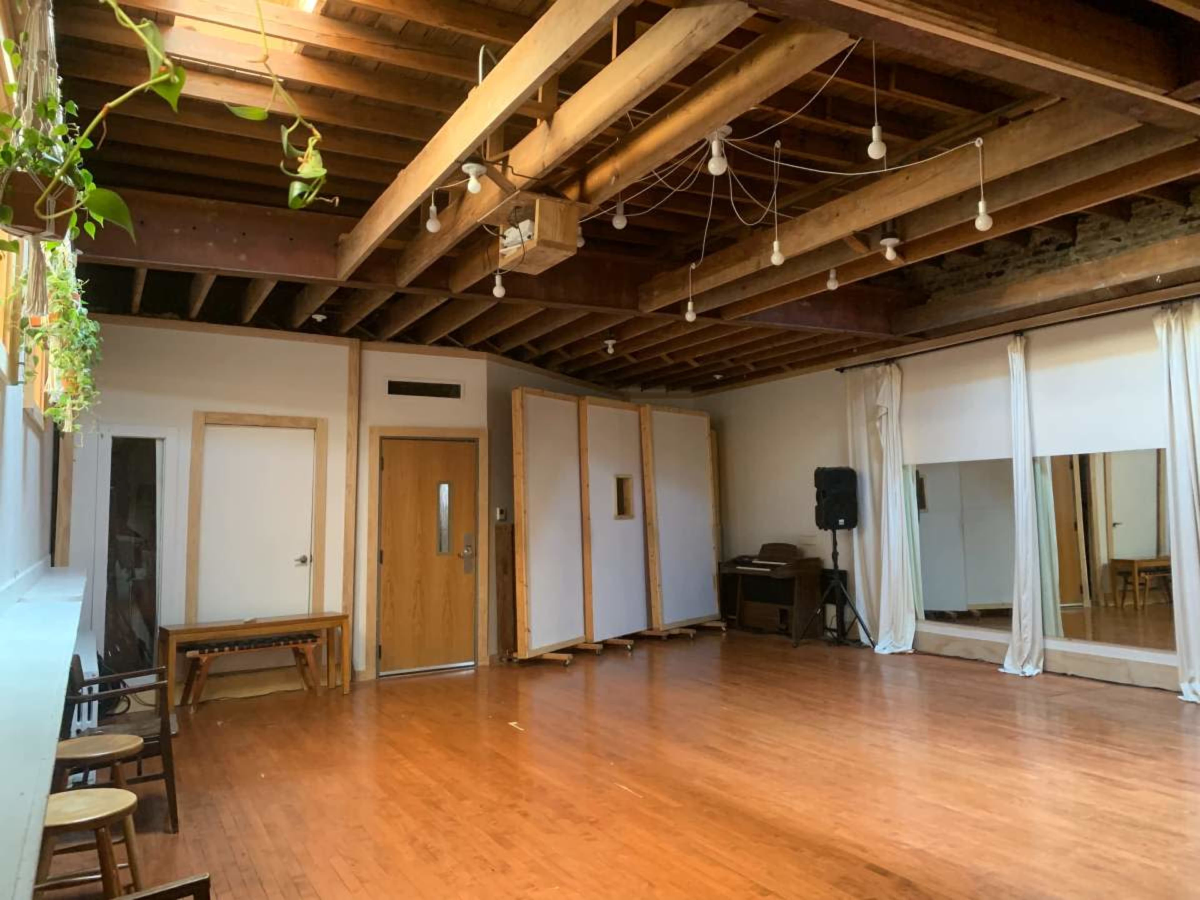 The image shows a spacious room with hardwood floors, exposed wooden beams, mirrors, and bright overhead lights.