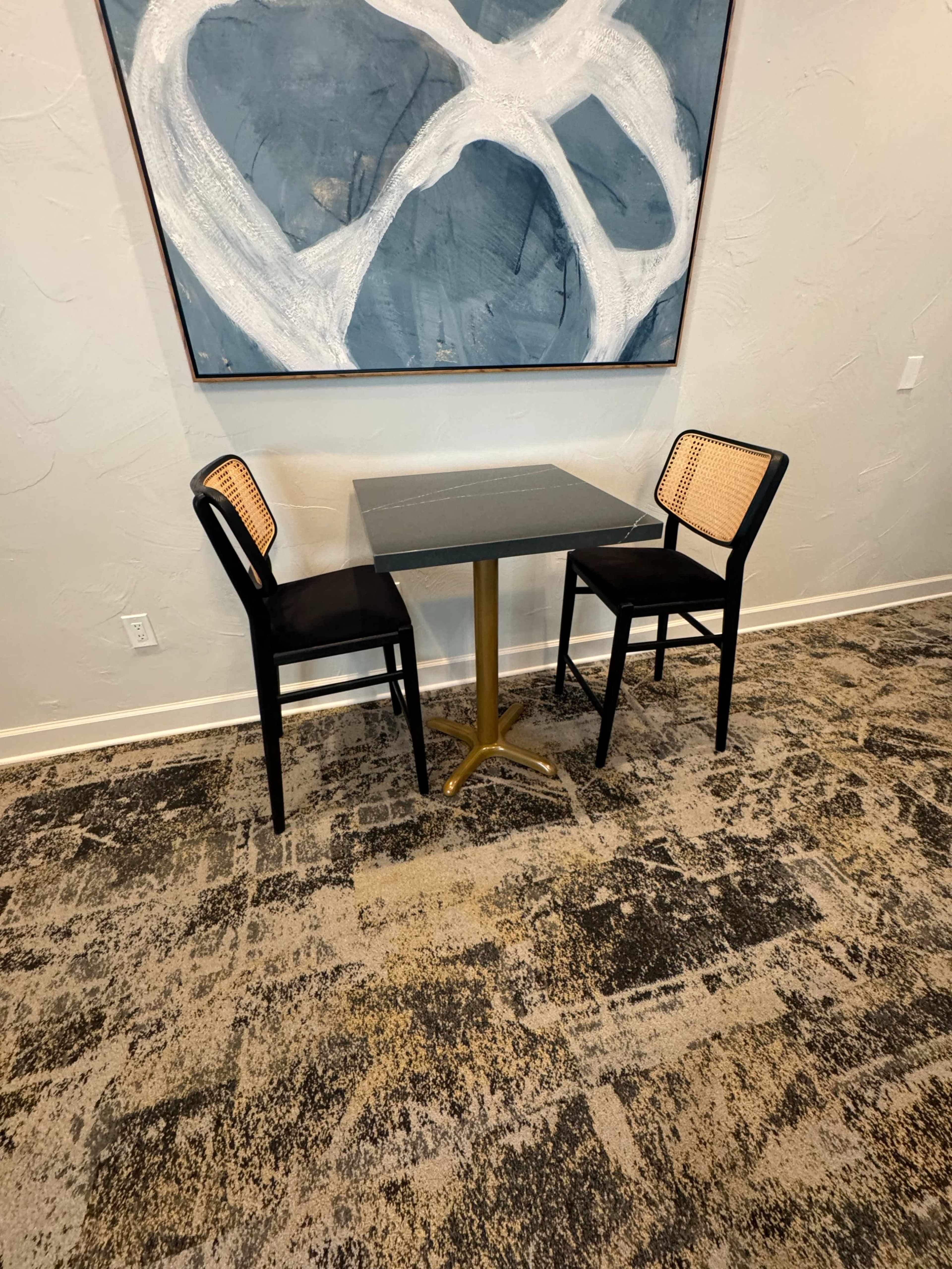 A small dining table with two black chairs featuring cane seats is set against a wall adorned with abstract artwork.