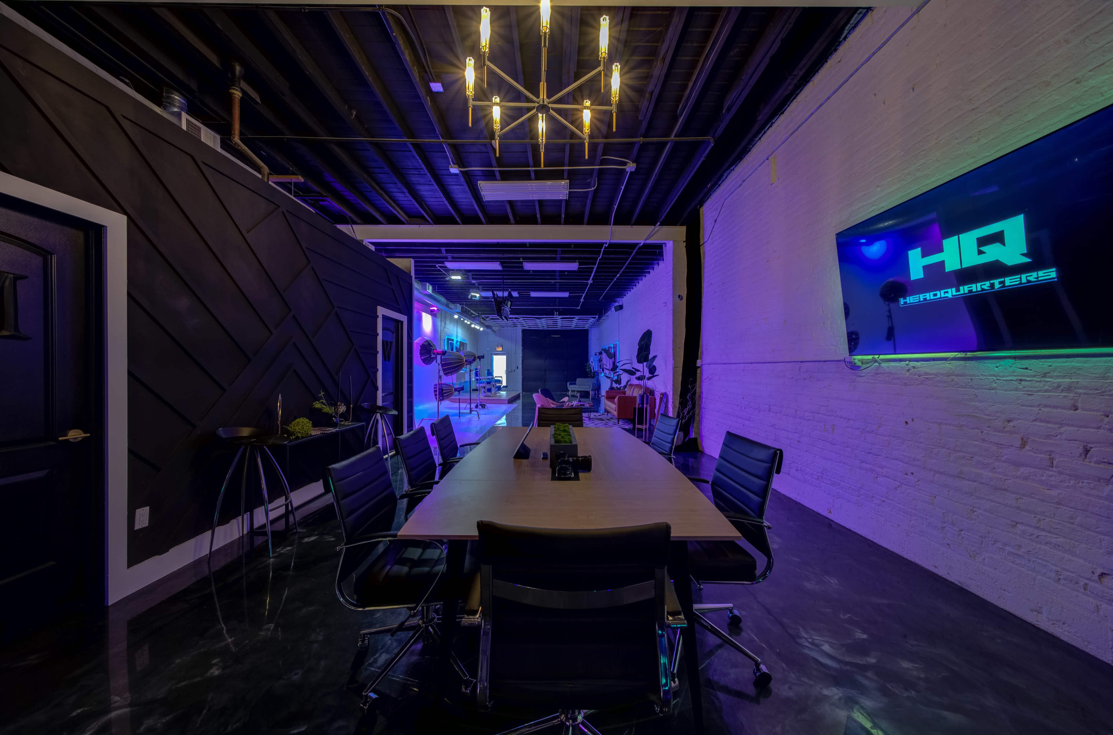 A modern conference room features a large table surrounded by black chairs, illuminated by a decorative chandelier and neon lighting on the walls.