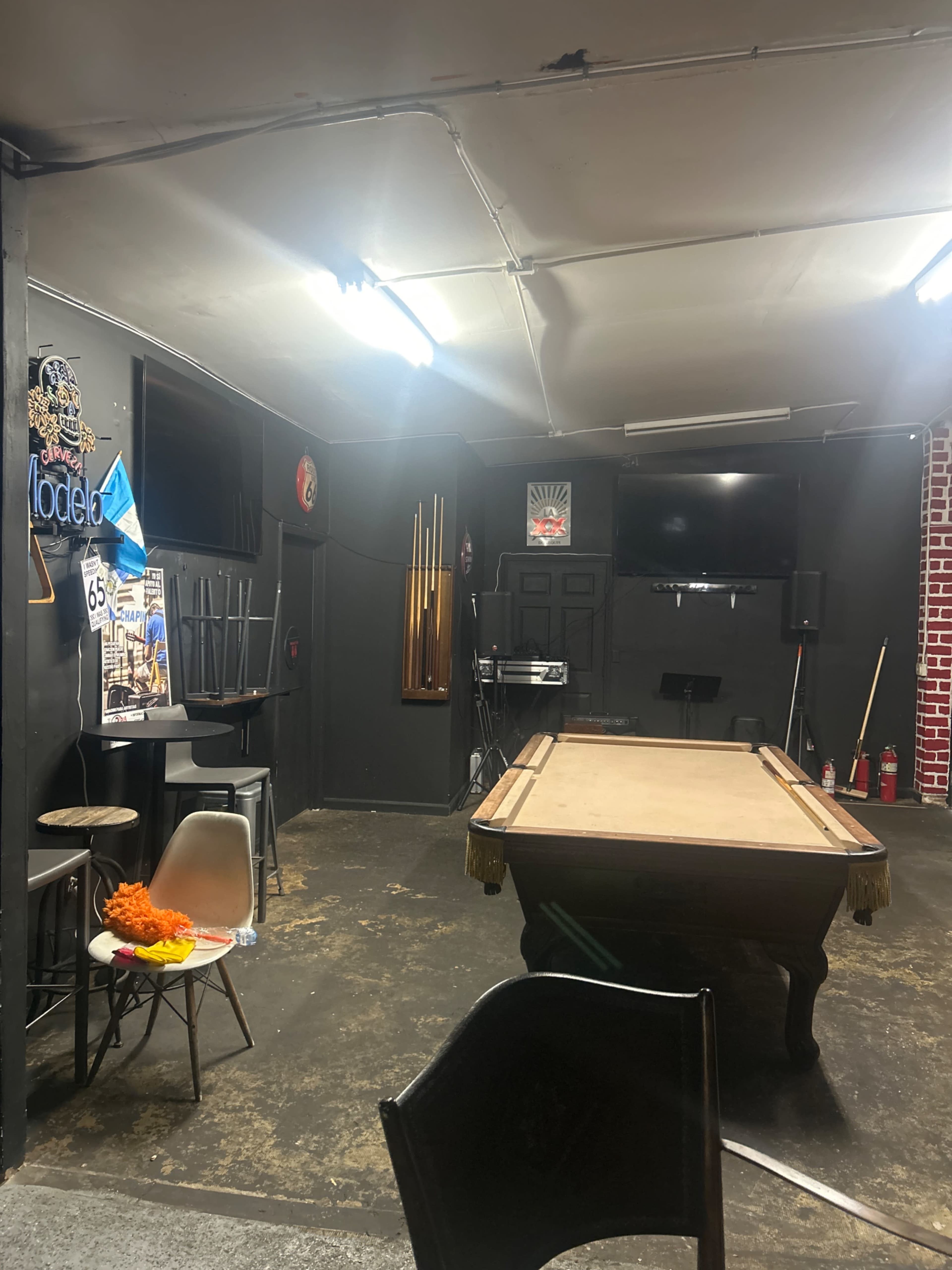 The image shows a dimly lit room with a pool table in the center, surrounded by chairs and featuring various decorations on the walls, including signs and musical equipment.