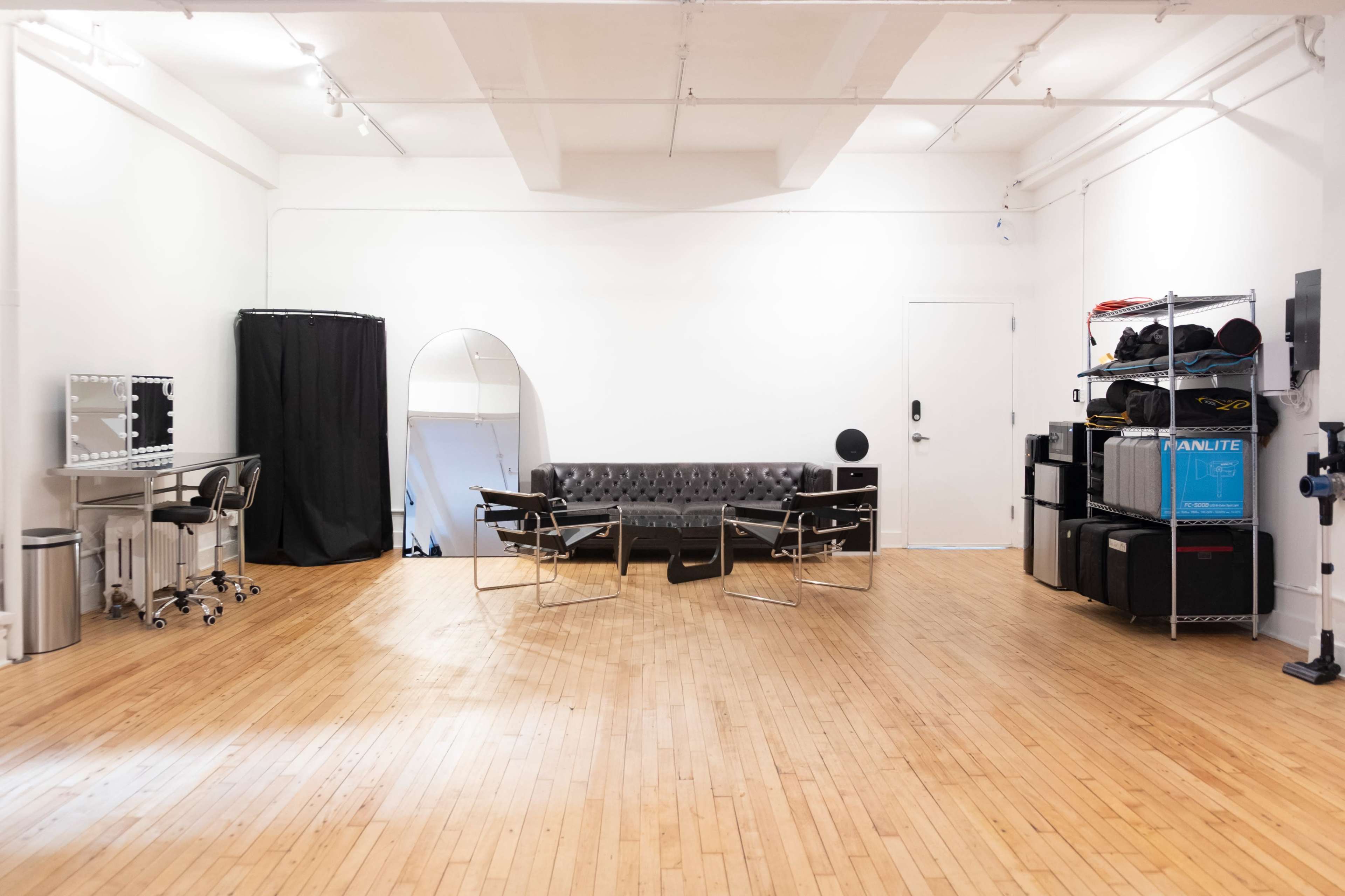 The image shows a spacious, well-lit studio with wooden flooring, featuring a black leather couch, two modern chairs, and various equipment neatly organized on shelves.