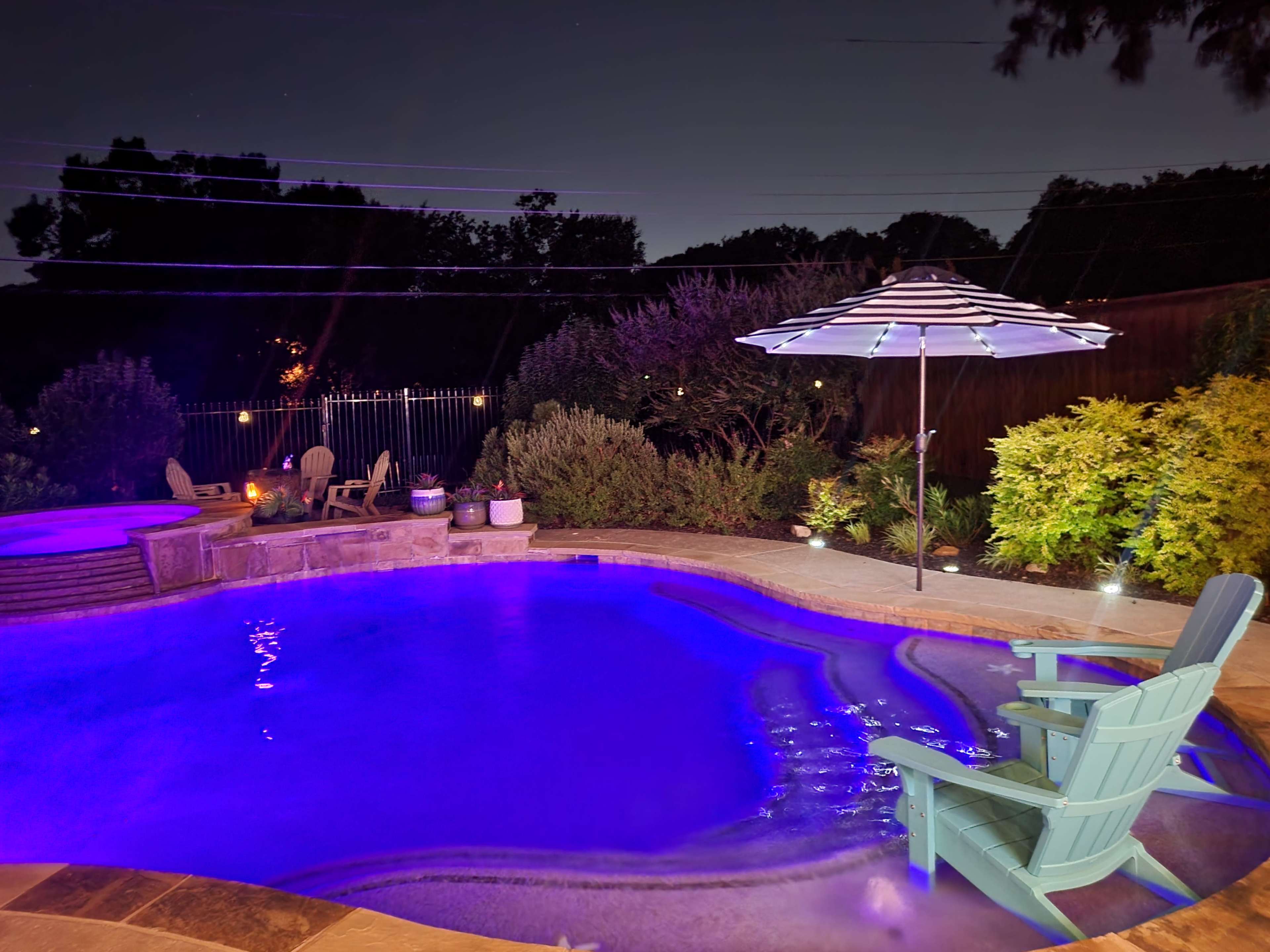 A illuminated swimming pool at night, featuring a lounge chair and an umbrella beside it, surrounded by landscaped greenery.