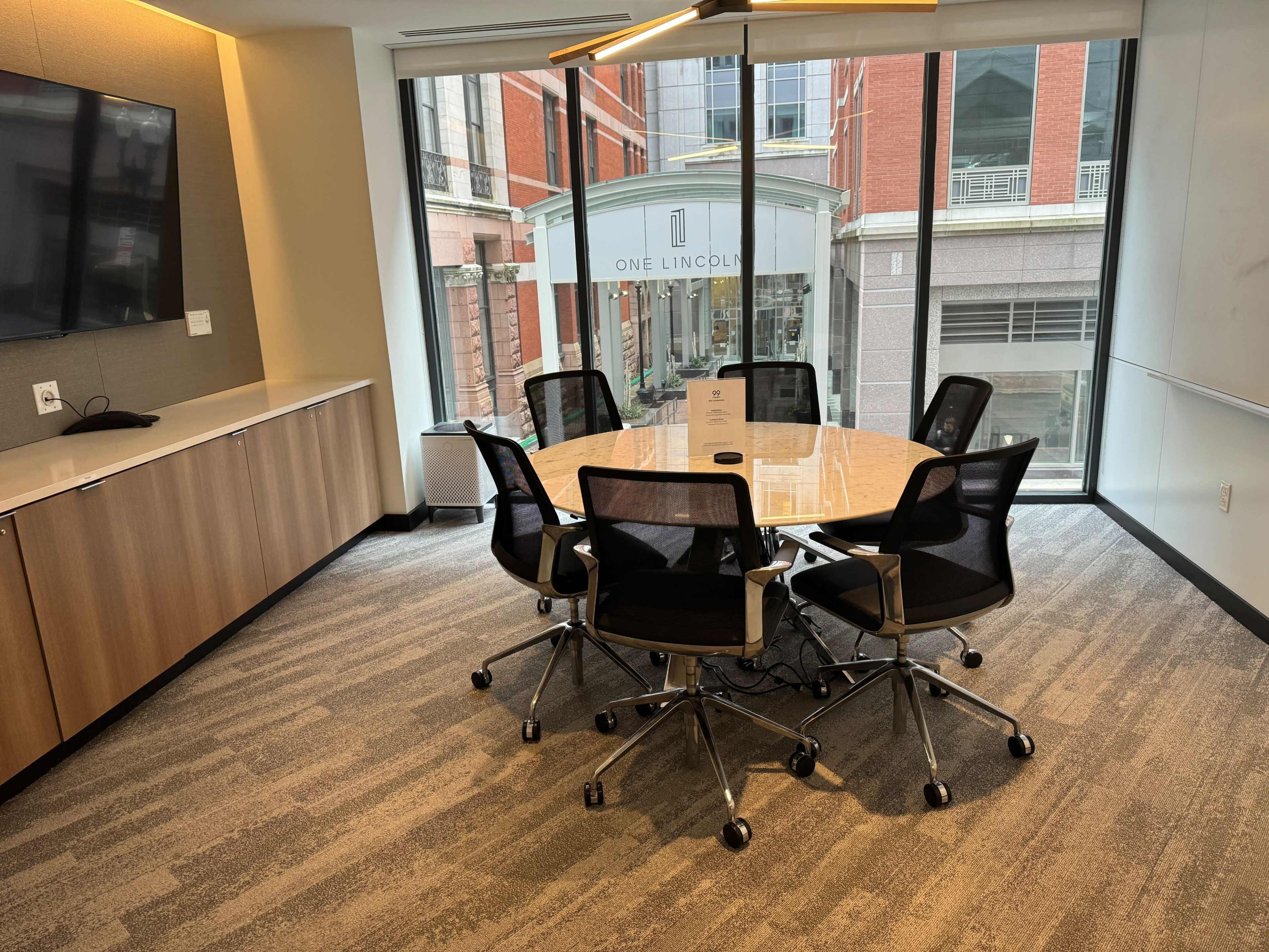 A modern conference room features a large circular table surrounded by black office chairs, with a view of a building labeled "ONE LINCOLN" through the window.