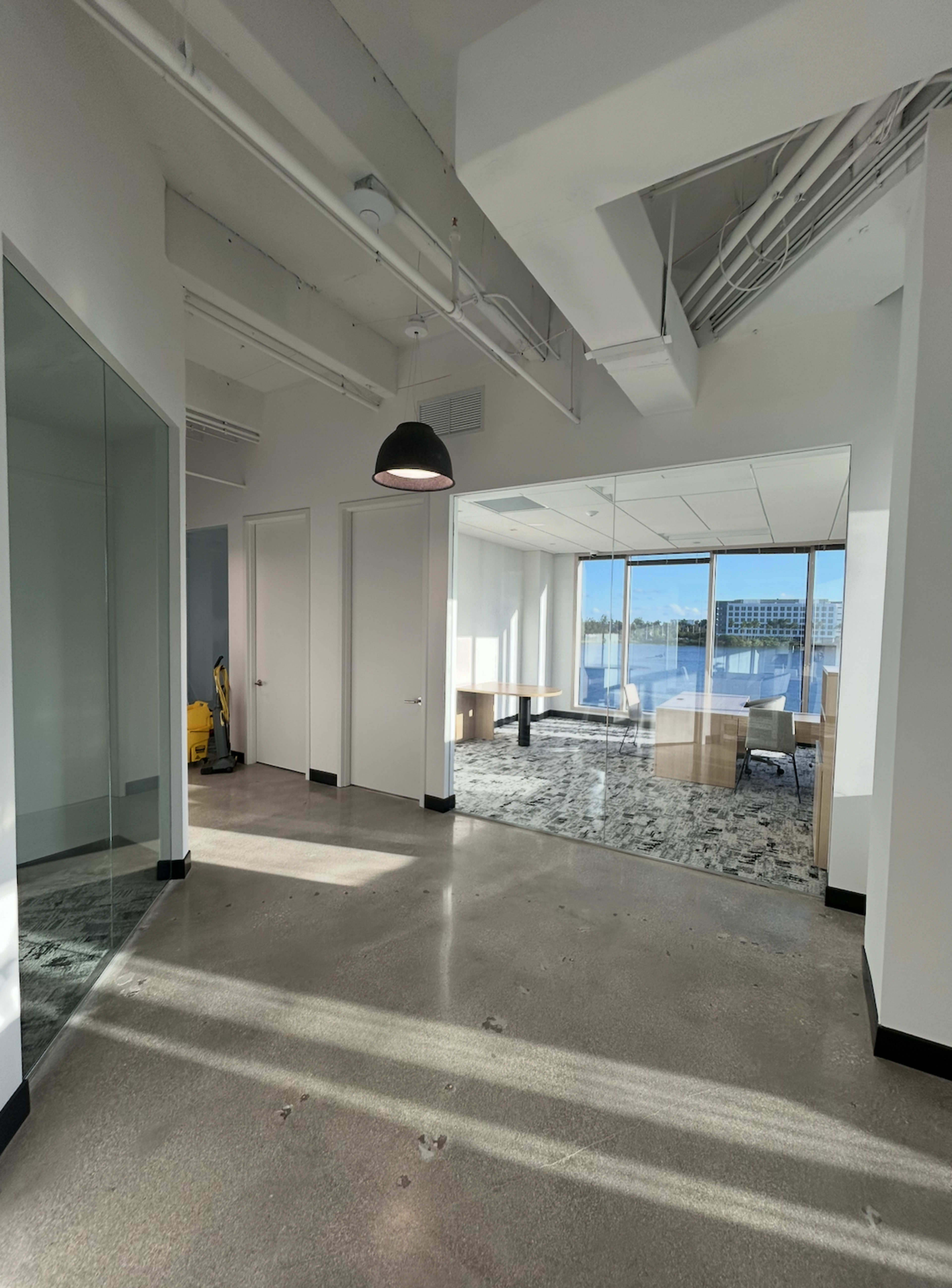 The image shows a modern office layout with glass walls, a conference room, and polished concrete floors, illuminated by natural light from large windows.