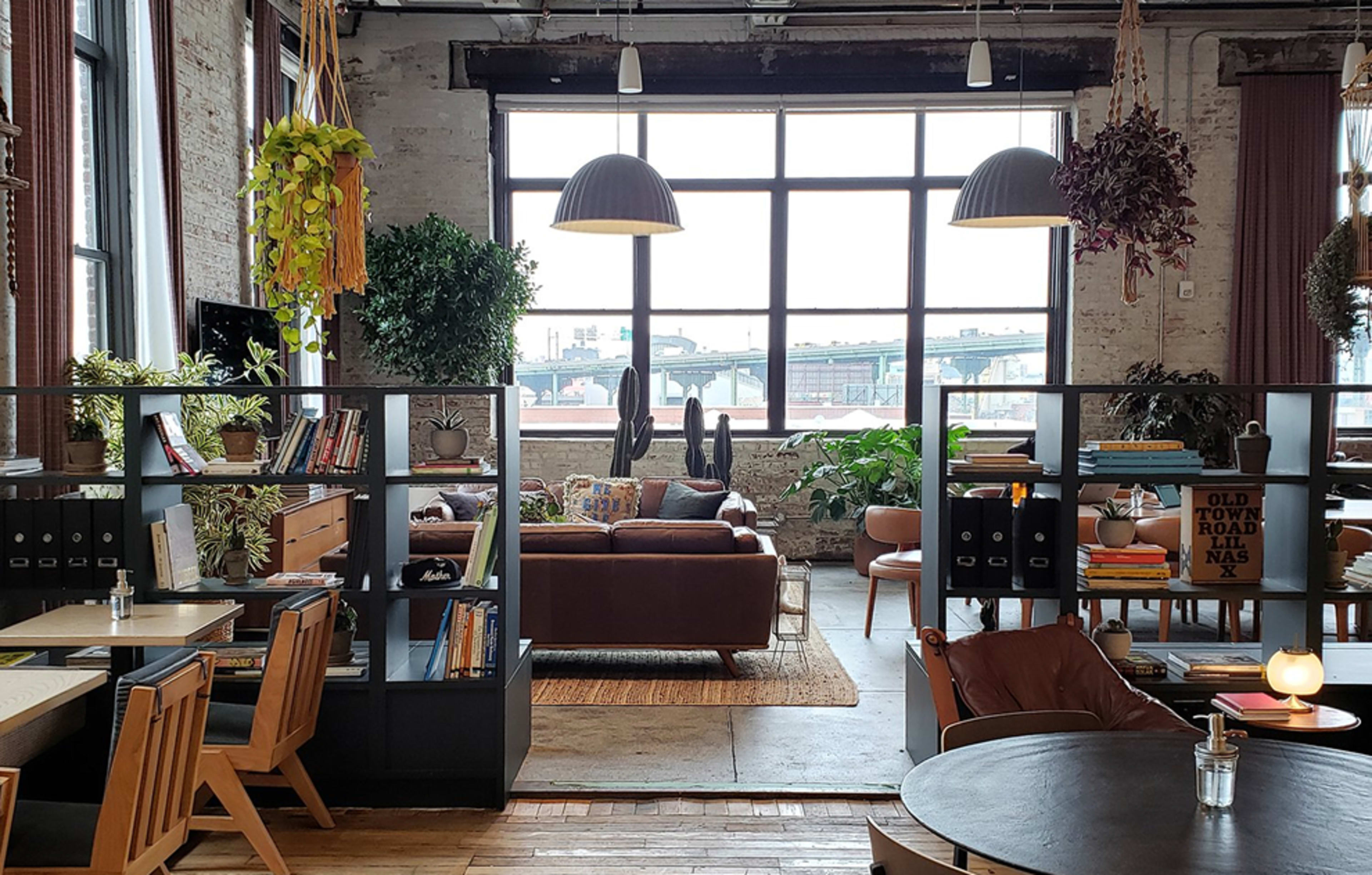 The image shows a stylish open-concept loft with large windows, a cozy seating area, and various plants arranged around shelves and furniture.