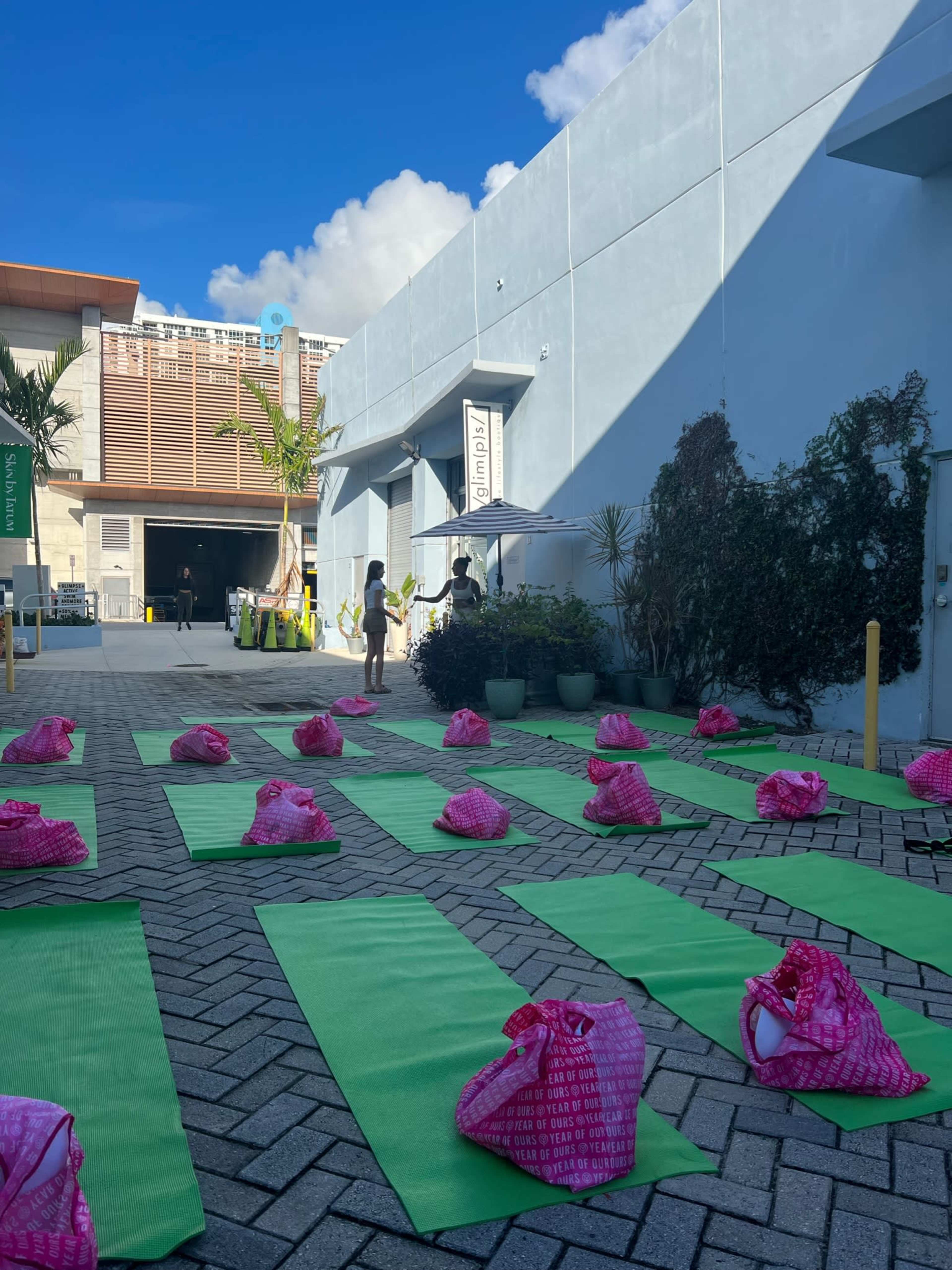 The image shows a sunny outdoor space set up for a yoga session, with green mats arranged neatly on the ground and pink bags placed at the top of each mat.