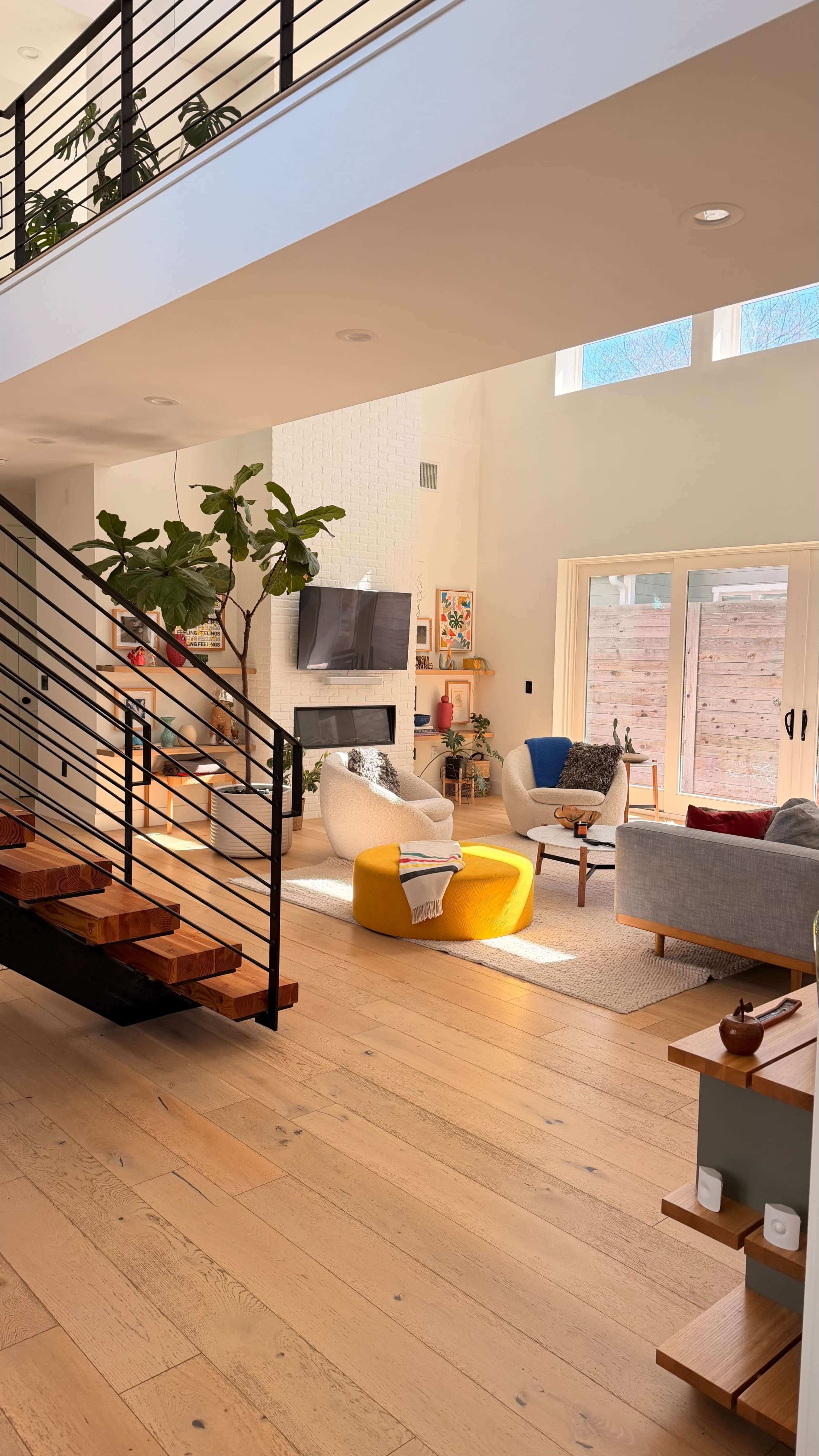 The image depicts a bright and spacious modern living room with a staircase, a sectional sofa, a round yellow ottoman, and large windows overlooking a patio area.
