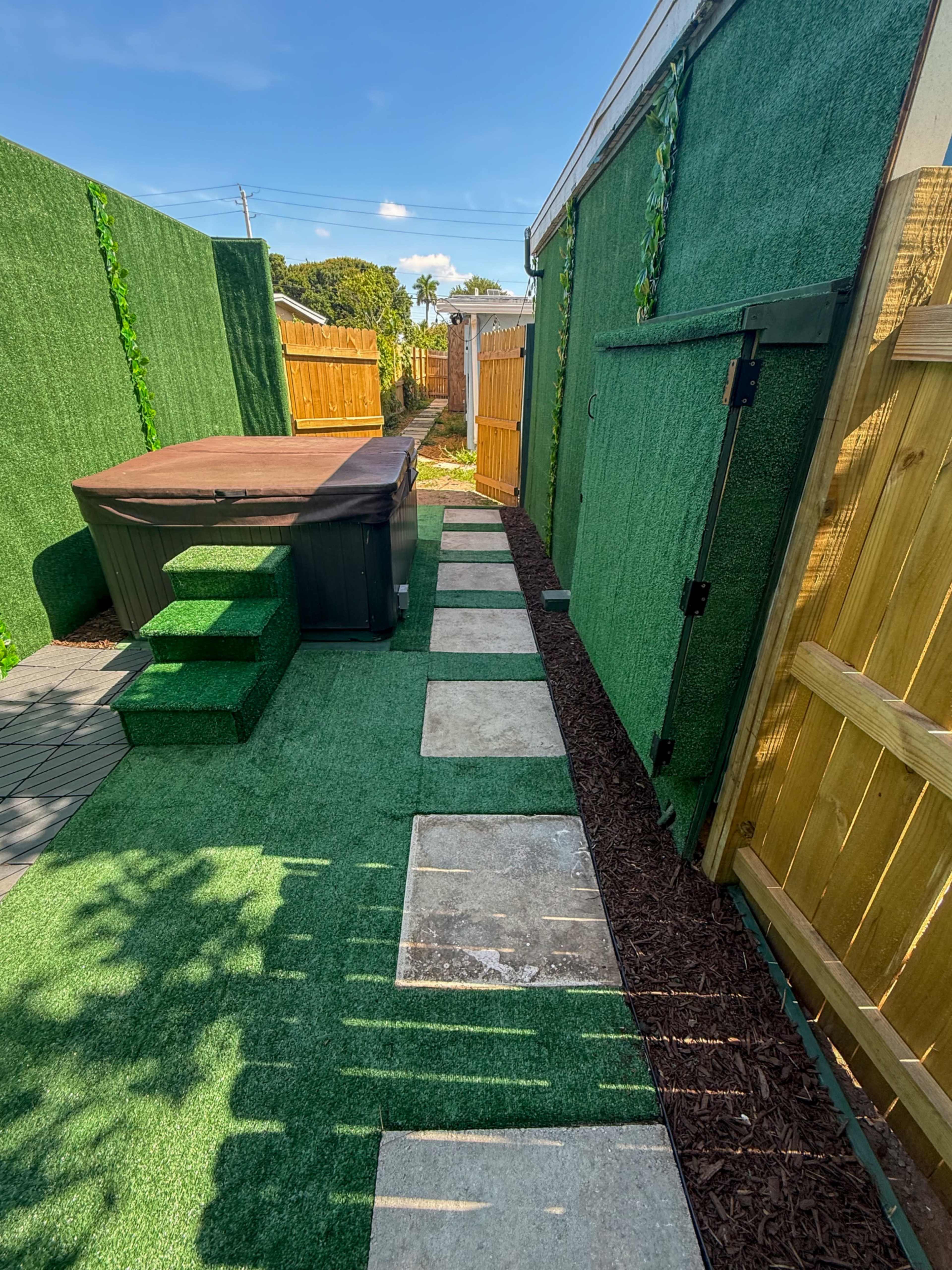 The image shows a landscaped outdoor area featuring a hot tub, stone pathway, artificial grass, and wooden fencing on either side.