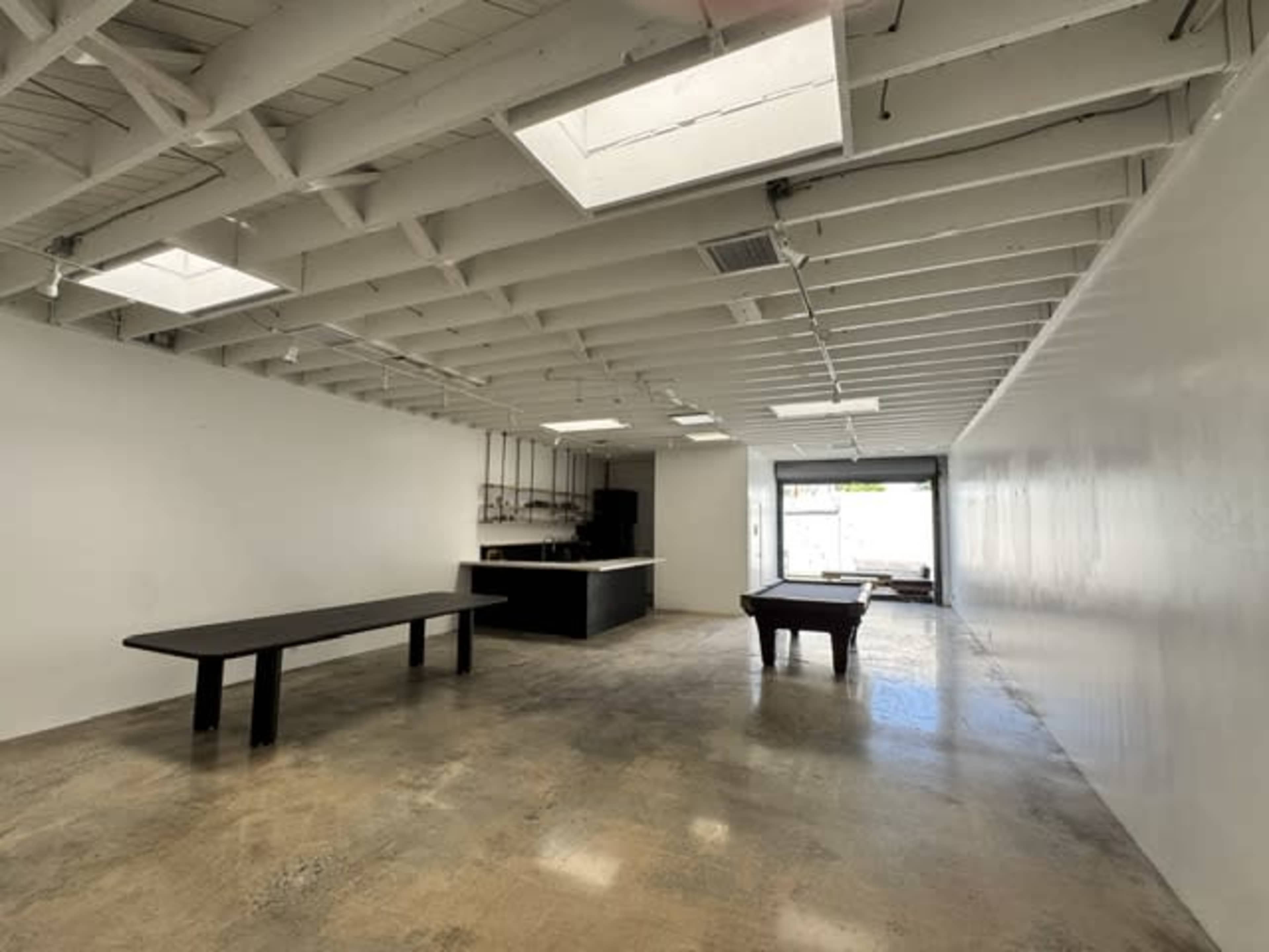 The image shows a spacious, empty room with a polished concrete floor, a large table, and a billiard table, illuminated by natural light from skylights and windows.