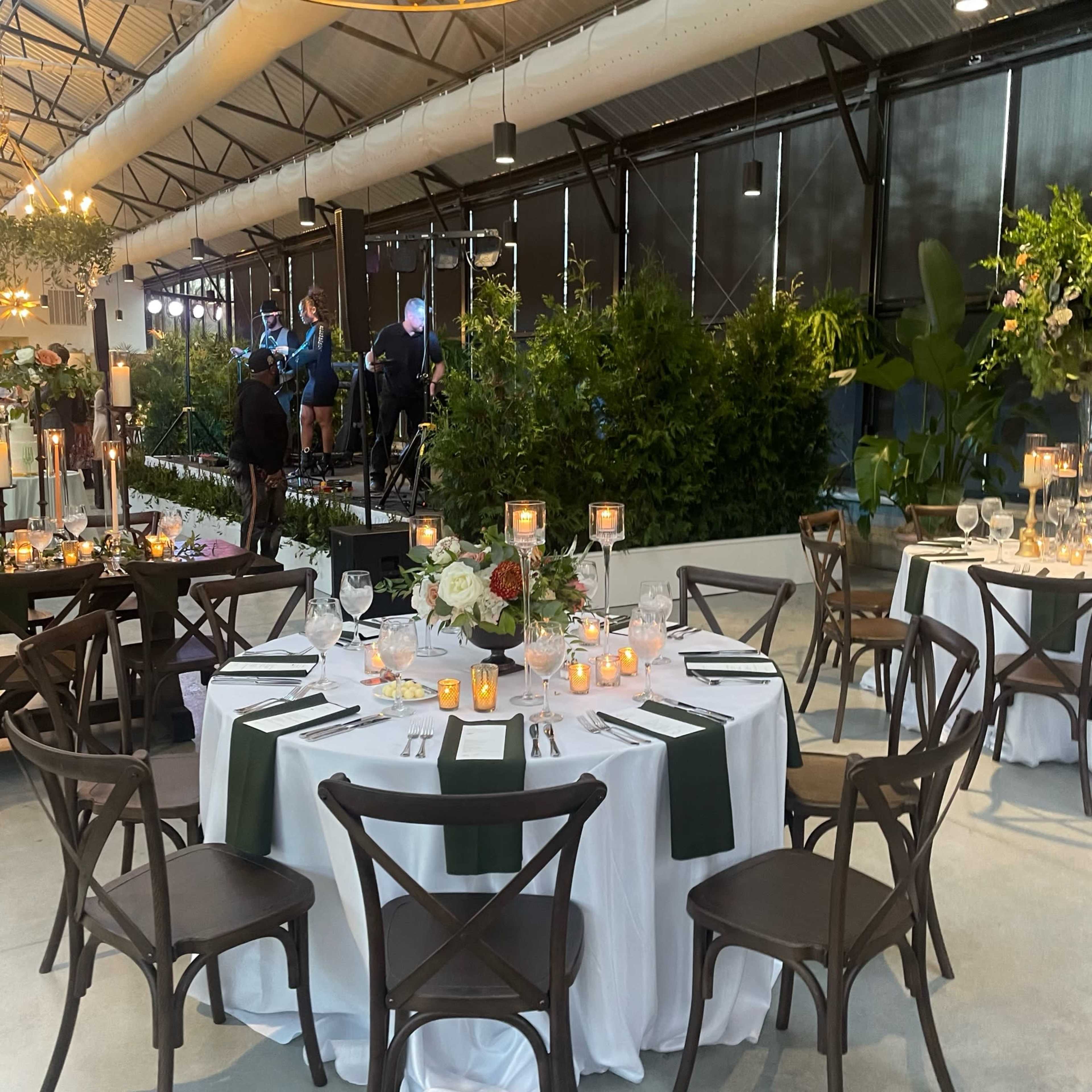 A beautifully set dining area features round tables with white tablecloths and green accents, surrounded by greenery, and a small stage in the background where musicians perform.