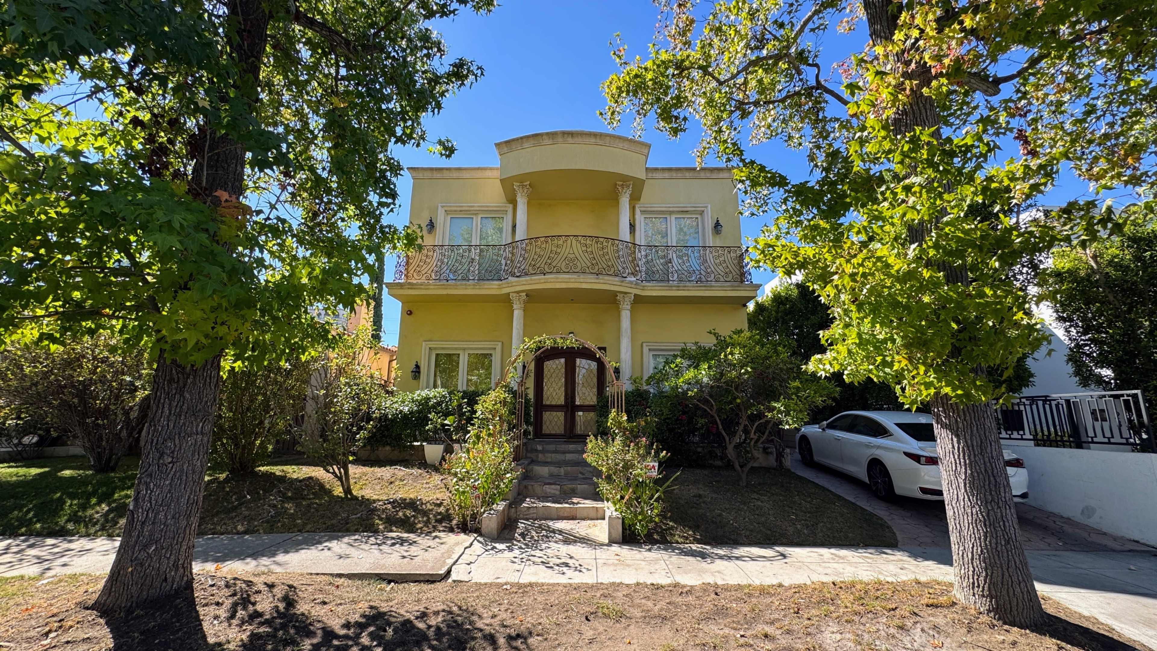 A two-story yellow house with a balcony is surrounded by greenery and has a white car parked in front.