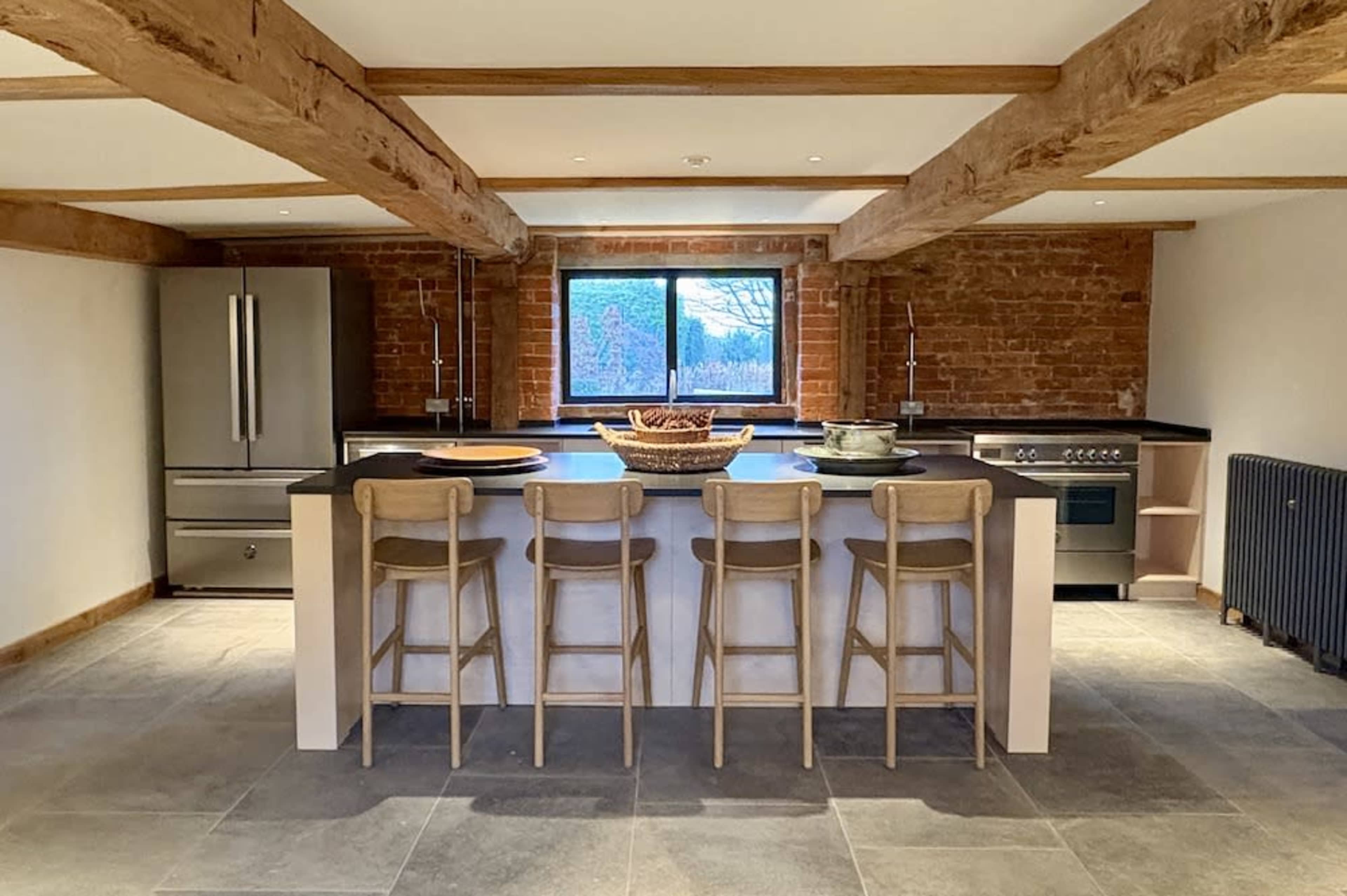 The image shows a modern kitchen featuring a large island with five wooden stools, stainless steel appliances, and exposed brick walls.