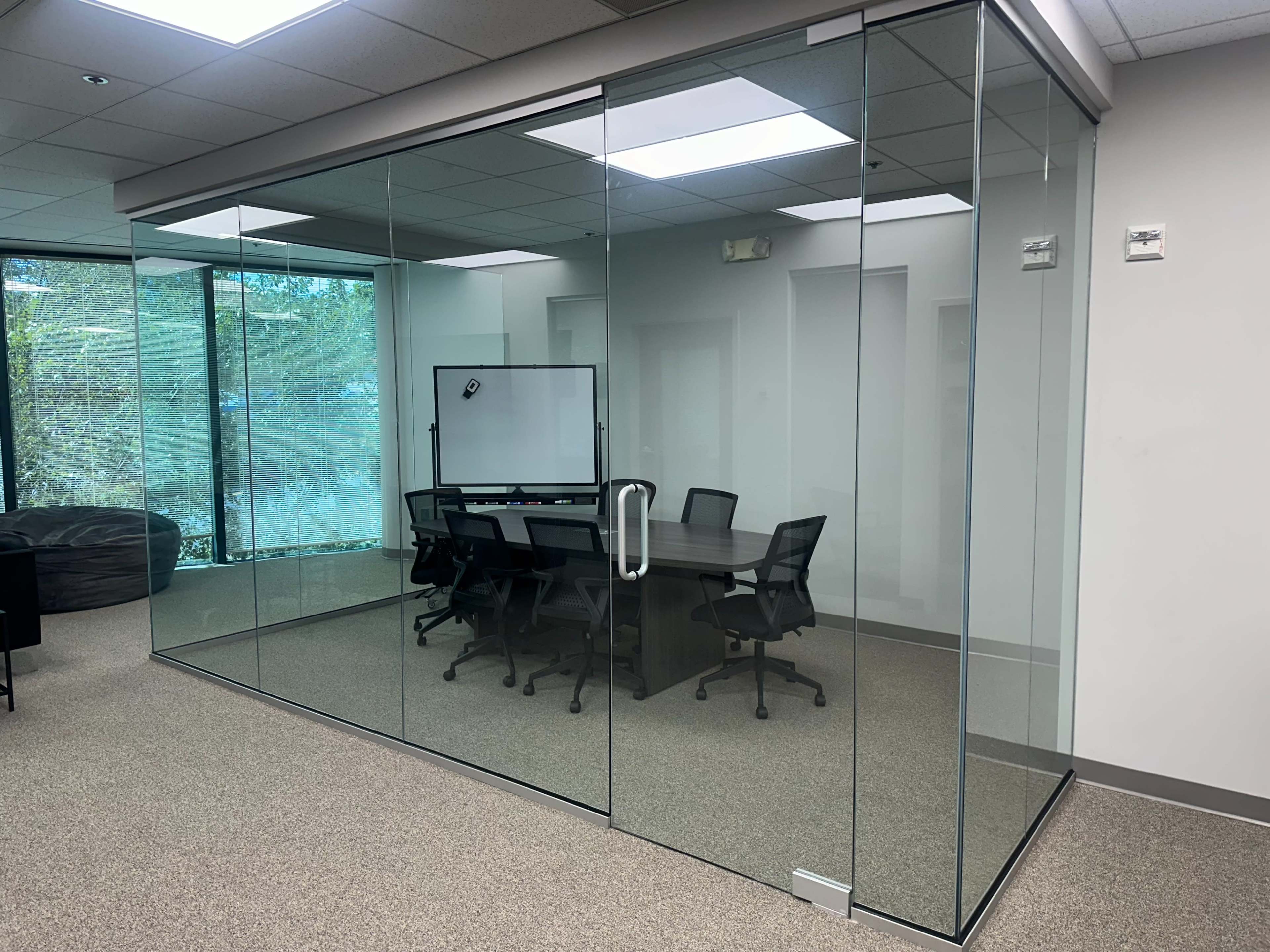 The image shows a modern conference room enclosed by glass walls, featuring a large table and several black chairs inside.