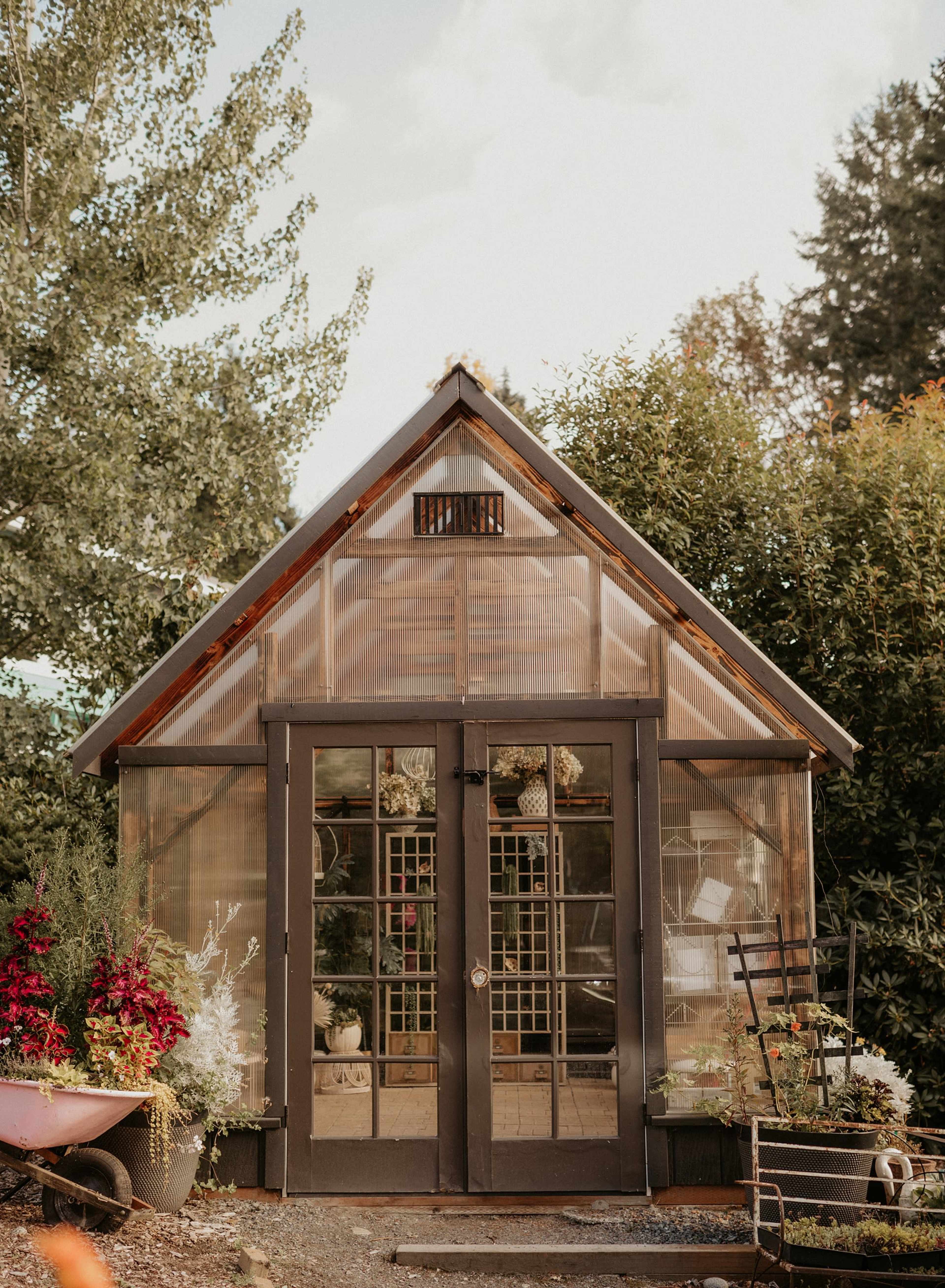 A wooden greenhouse with glass panels and double doors is surrounded by vibrant plants and flowers.