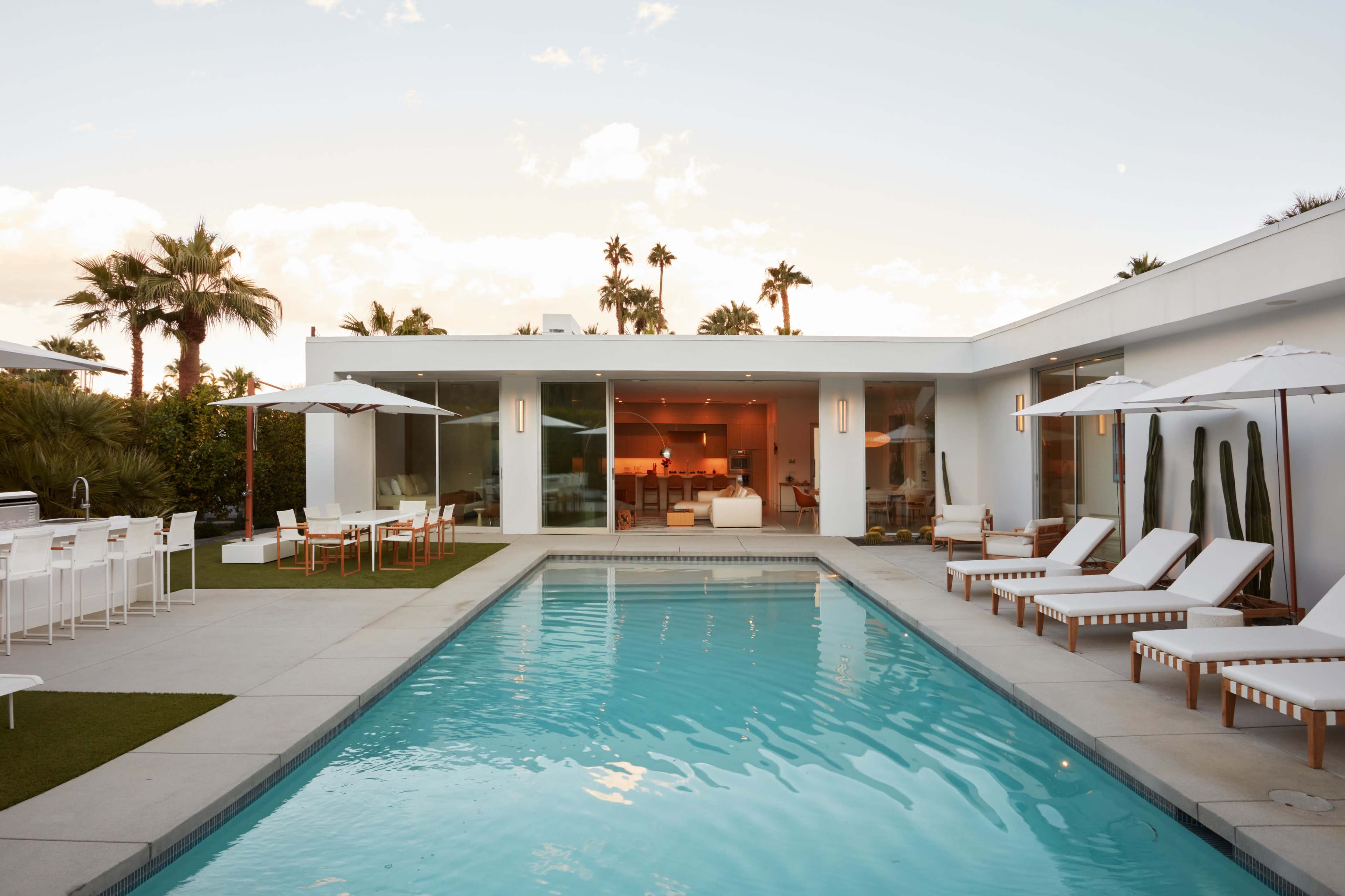 A modern home with large glass walls surrounds a rectangular swimming pool, featuring lounge chairs and patio seating under white umbrellas, set against a backdrop of palm trees.