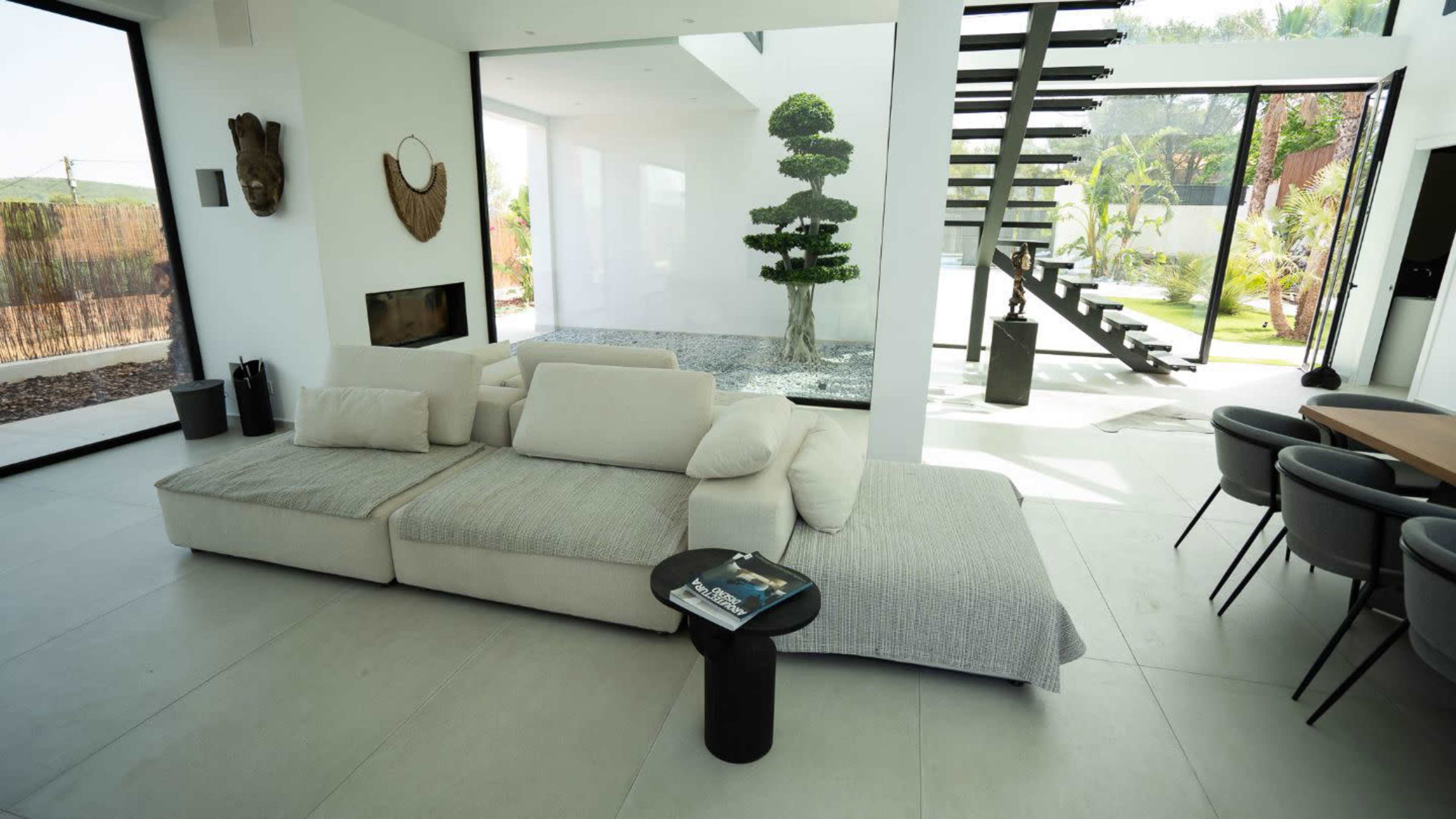 A modern living room features a sectional sofa, a small black side table, and large windows overlooking a garden and a staircase.
