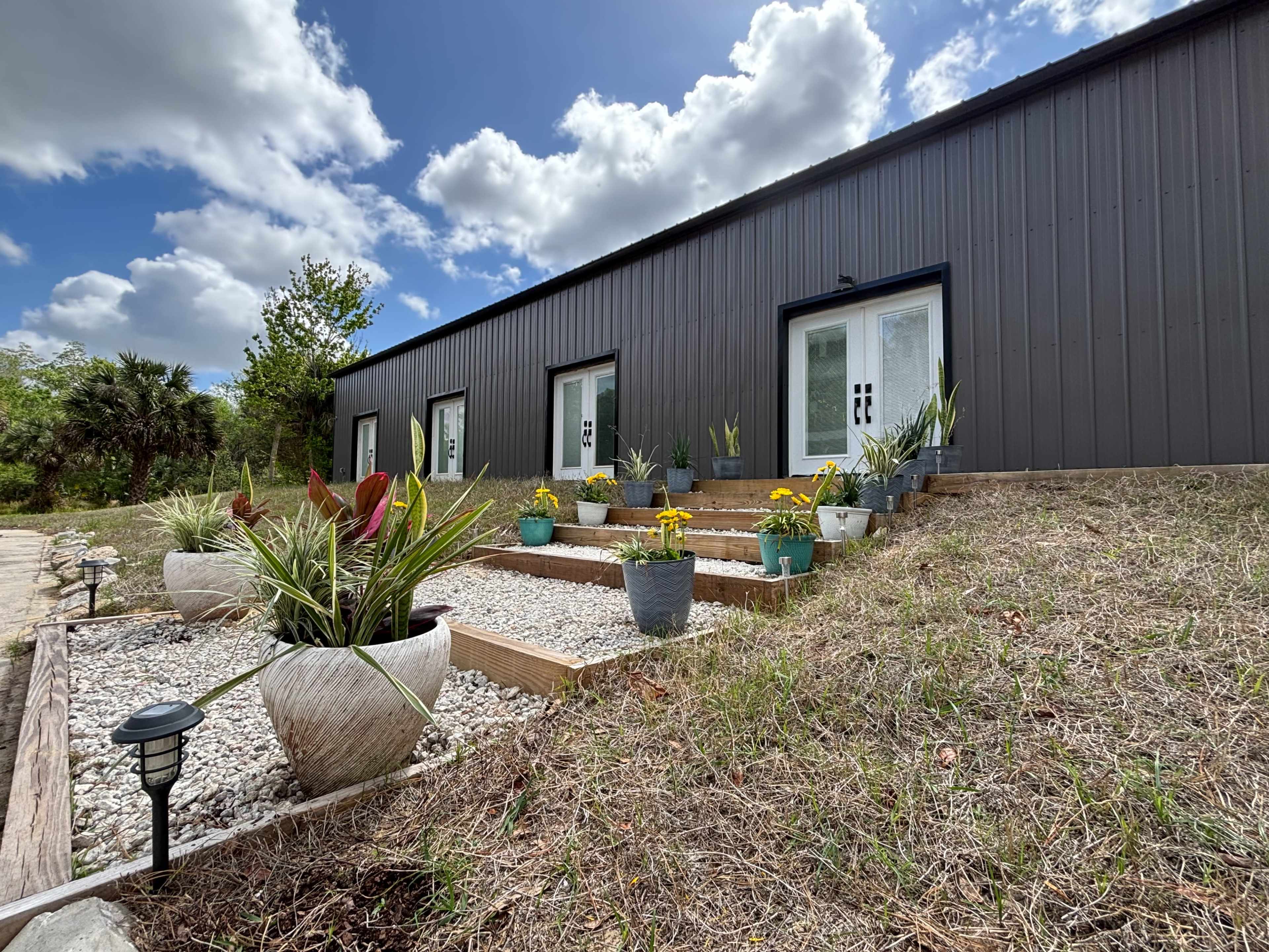 The image shows a modern building with a dark exterior, featuring a set of stairs adorned with potted plants and gravel landscaping under a partly cloudy sky.