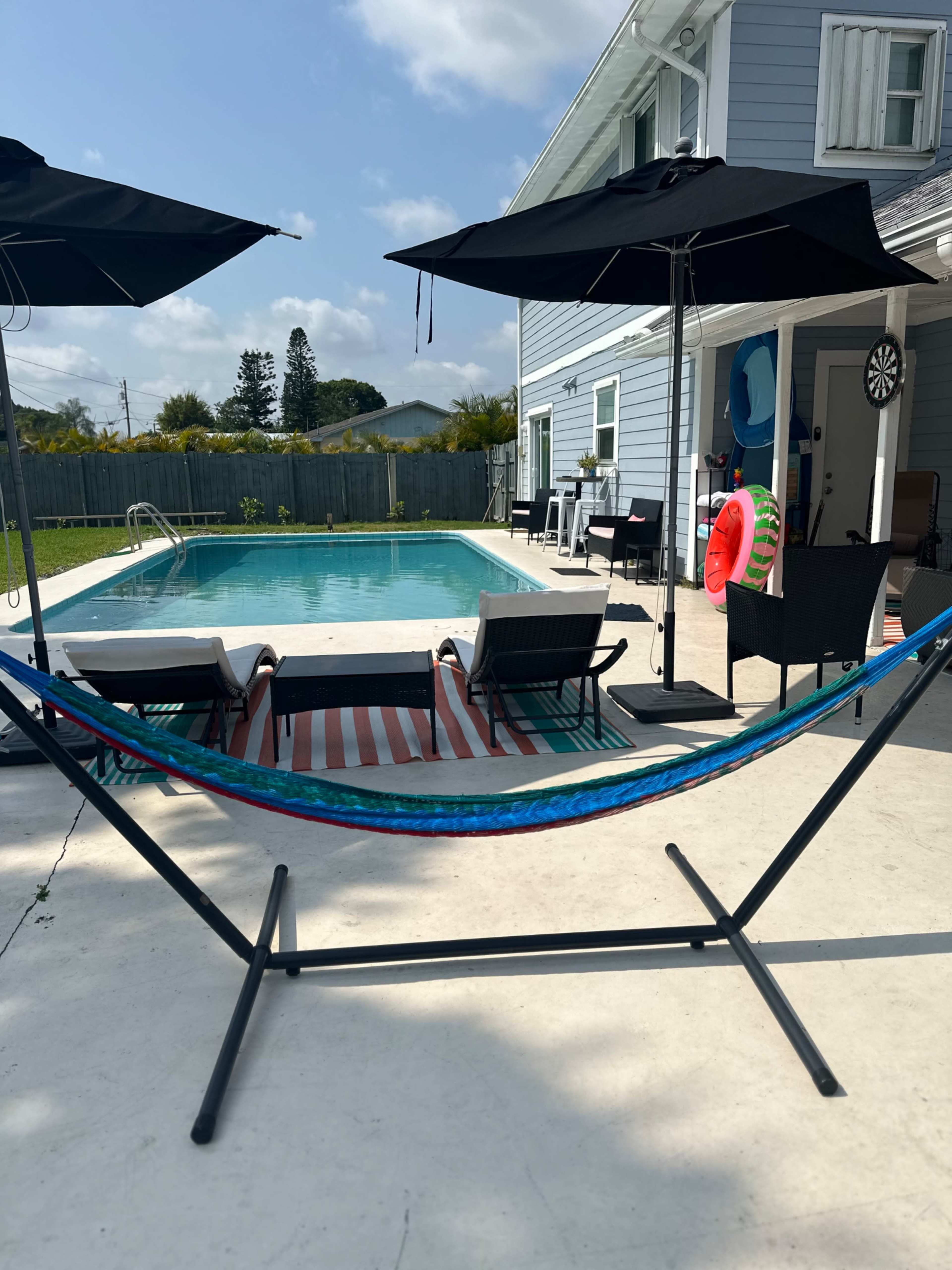 A hammock is set up near a swimming pool, surrounded by lounge chairs and patio umbrellas at a residential property.