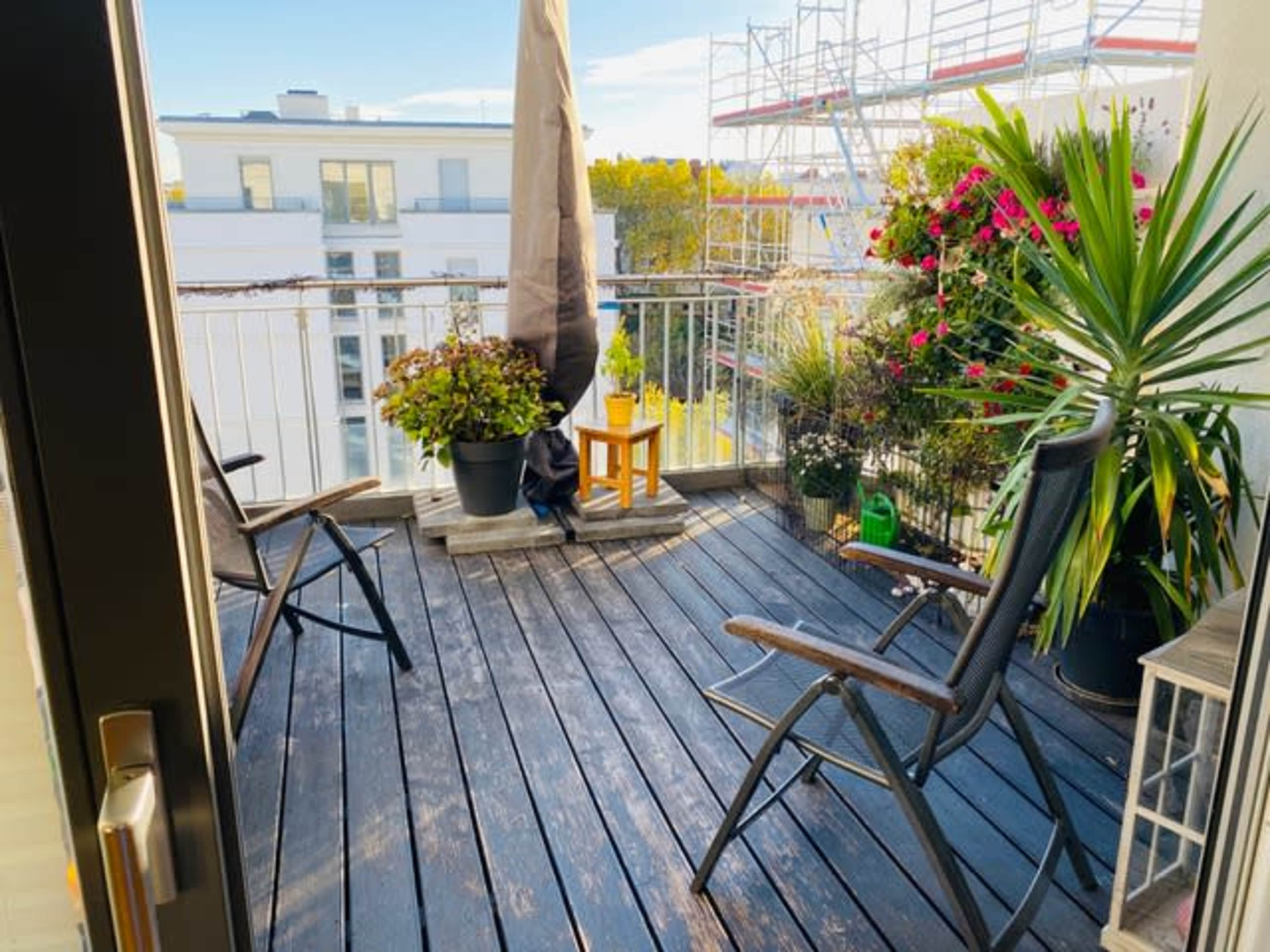 A small balcony features two chairs, potted plants, and a wooden table under a large umbrella.