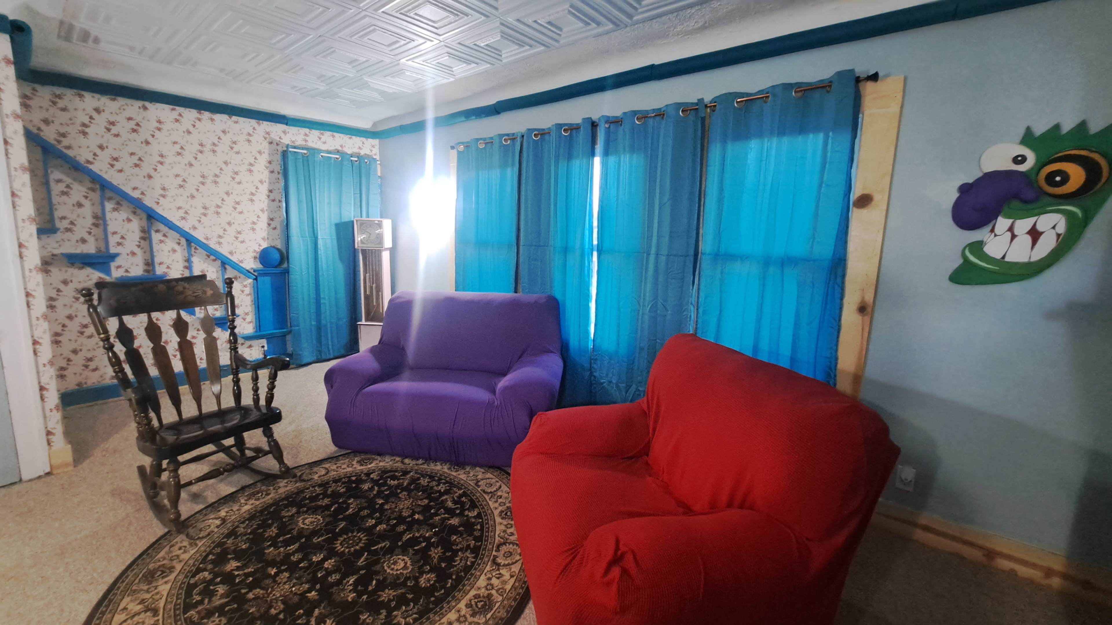 A brightly lit living room features a purple chair, a red chair, a patterned area rug, and blue curtains against a floral wallpaper backdrop.