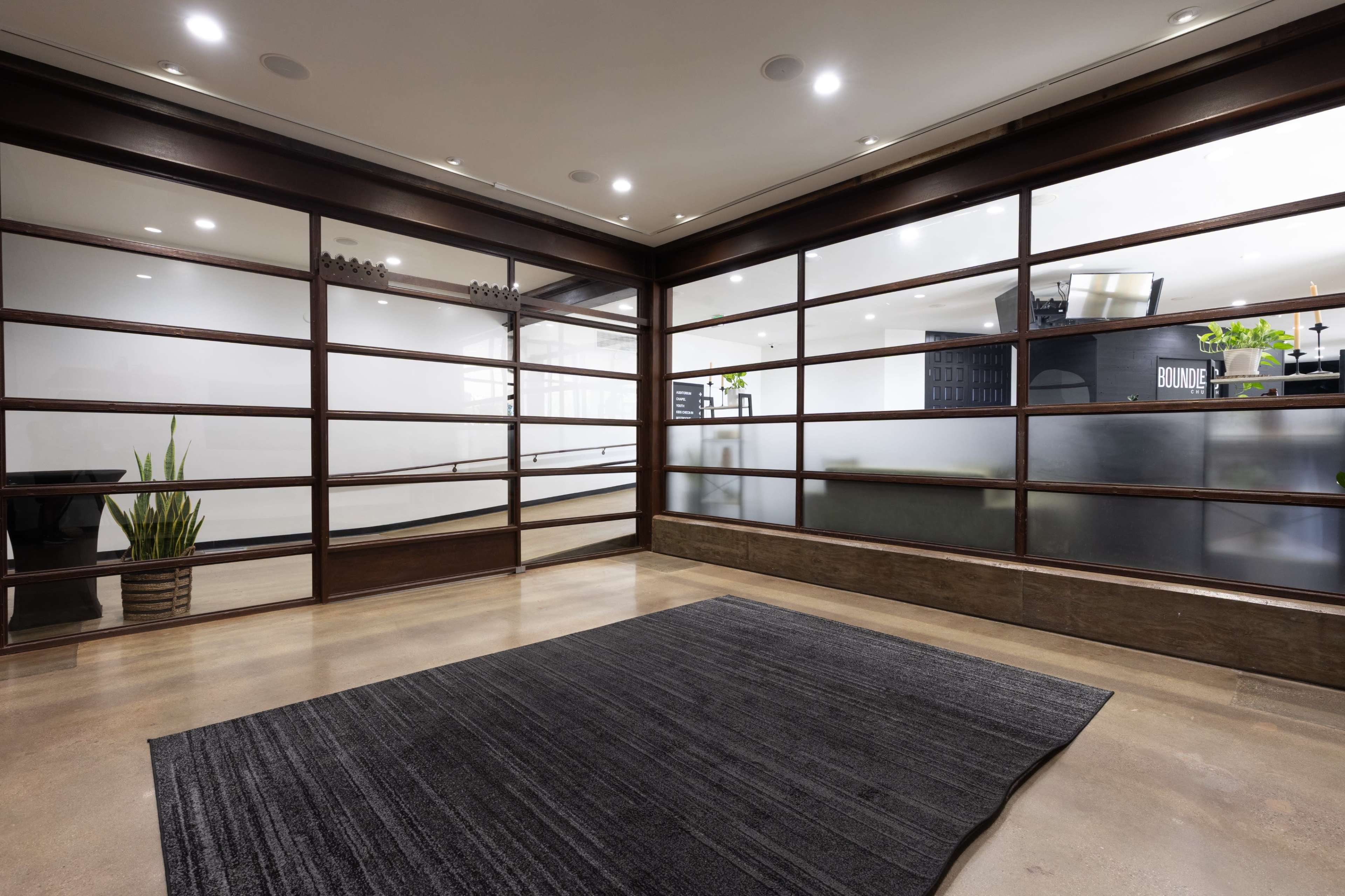 The image shows a modern, open office space with large glass walls, a concrete floor, and a dark area rug.