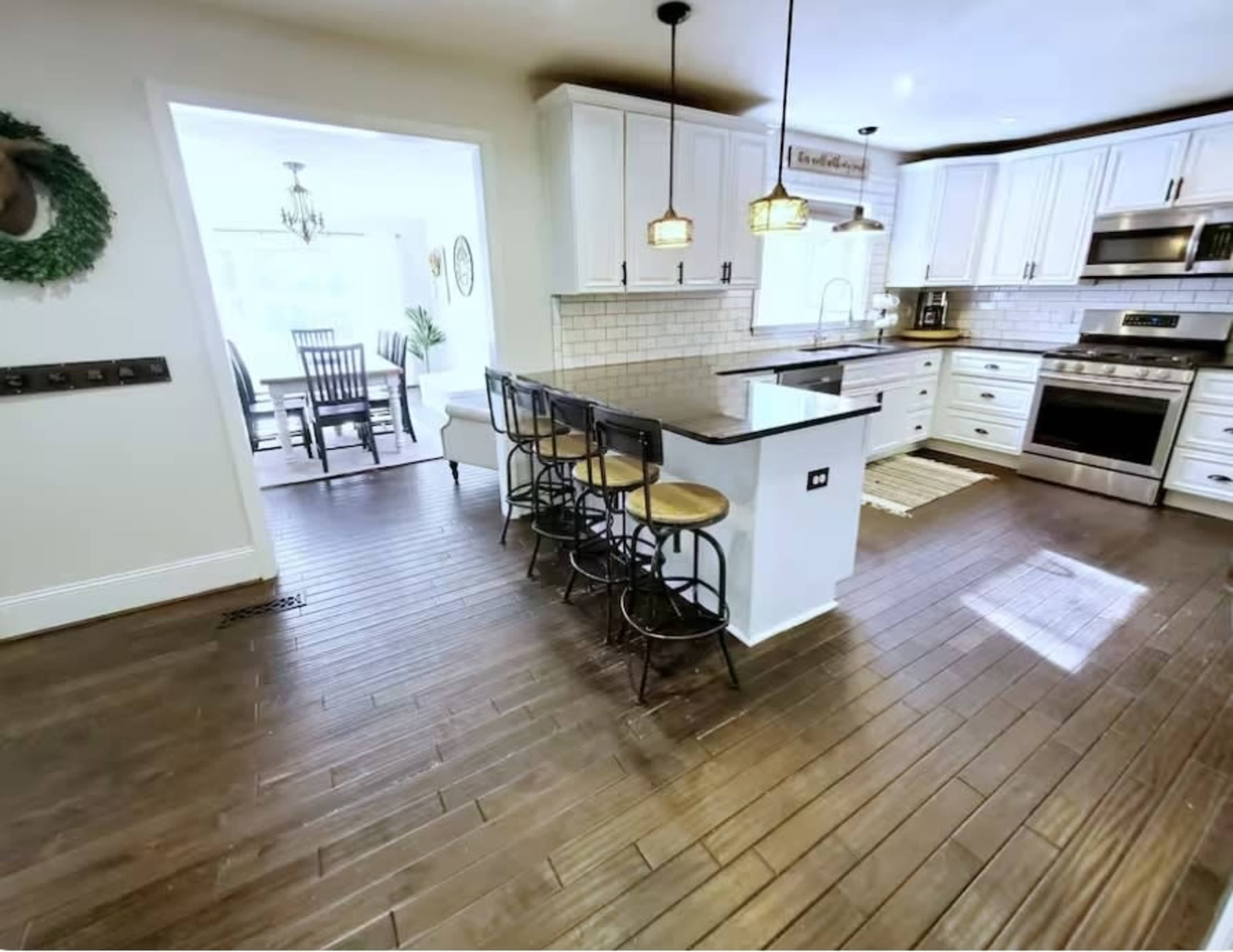 A modern kitchen features wooden flooring, white cabinets, a central island with bar stools, and a dining area visible in the background.