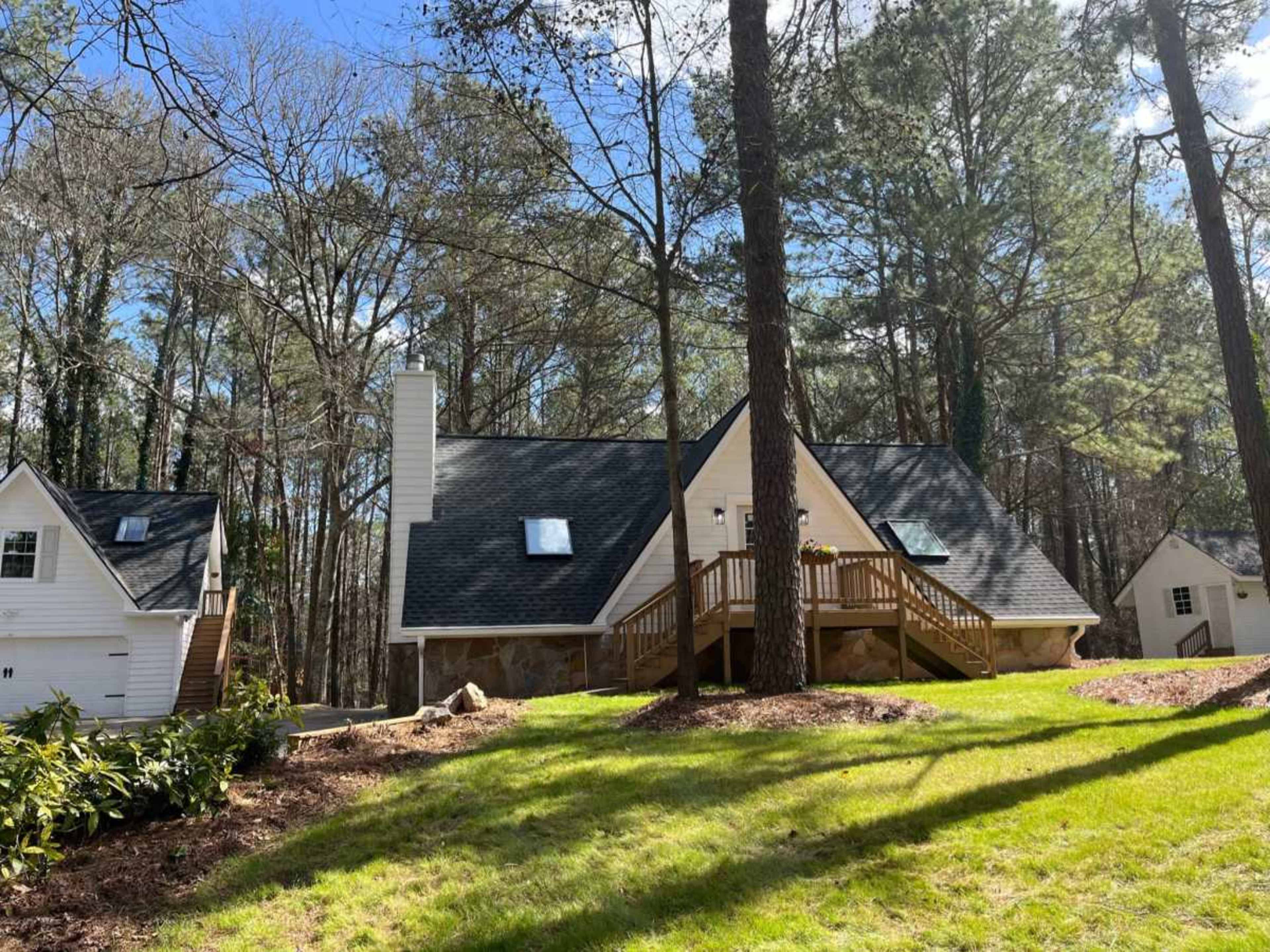A two-story house with a steep roof and wooden deck is surrounded by trees in a grassy area.