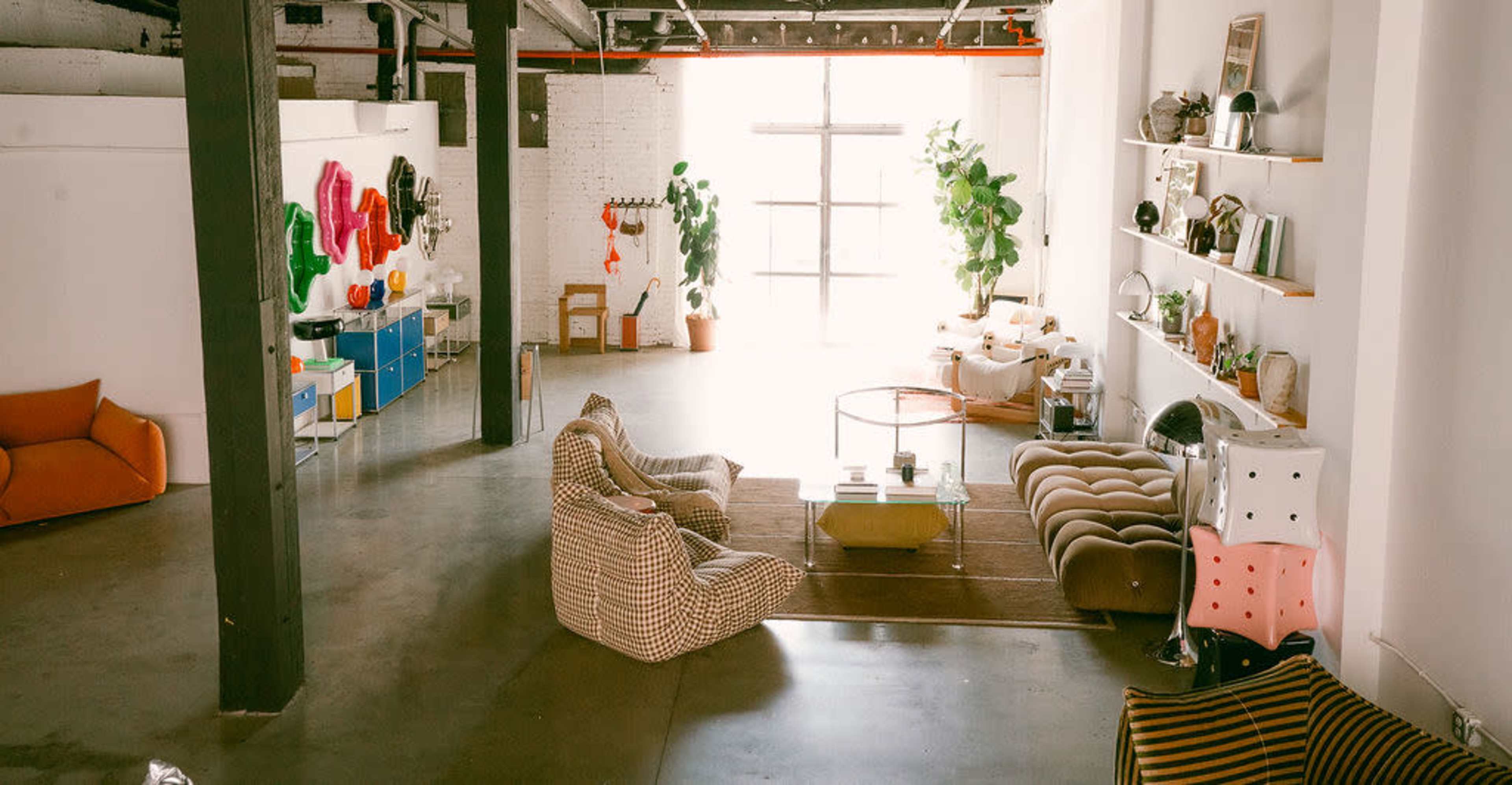 The image shows a spacious, modern loft with large windows, featuring various seating areas, decorative plants, and minimalist shelving units.