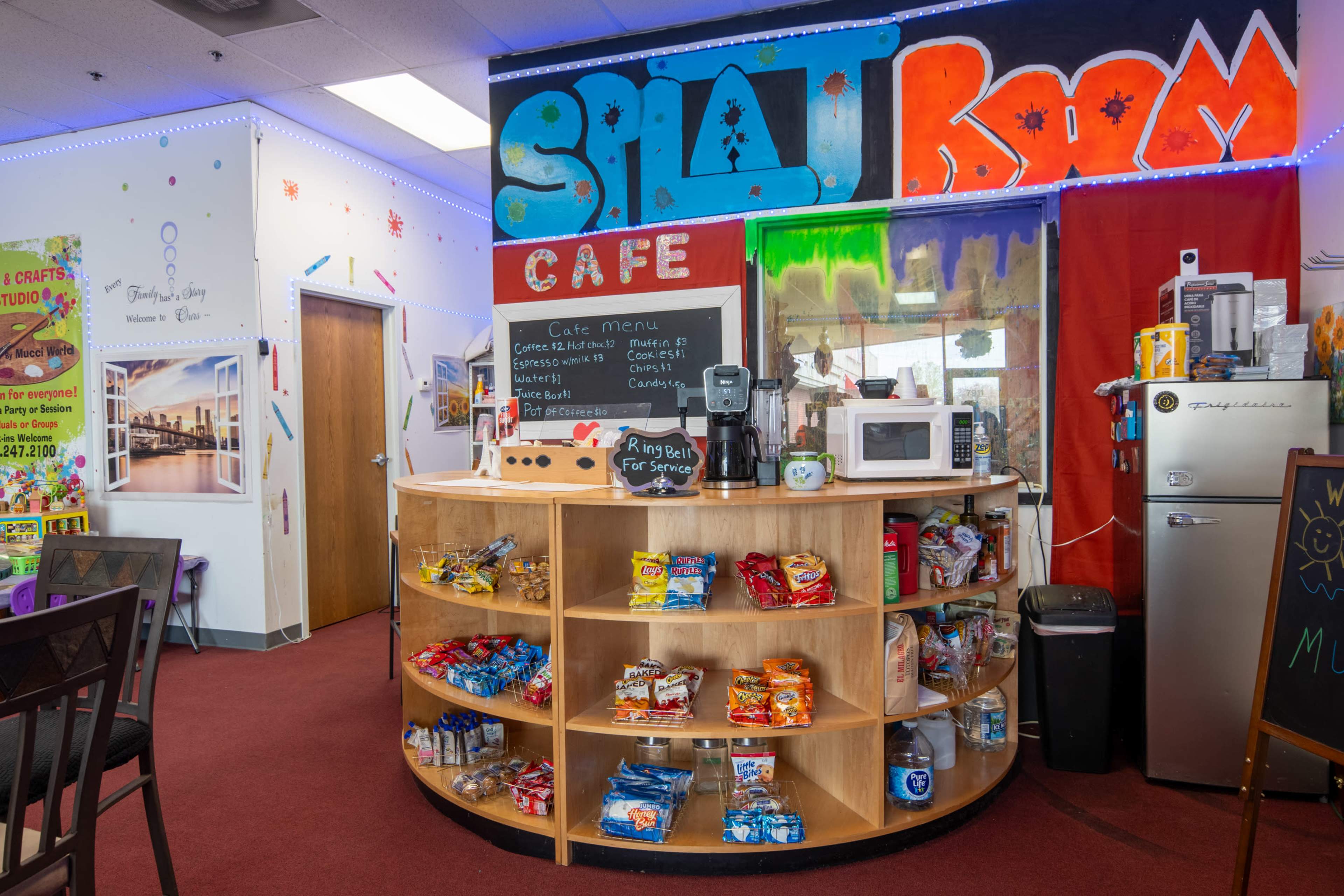A cafe area with a round wooden shelf displaying snacks and beverages, surrounded by colorful wall decor and a service counter.