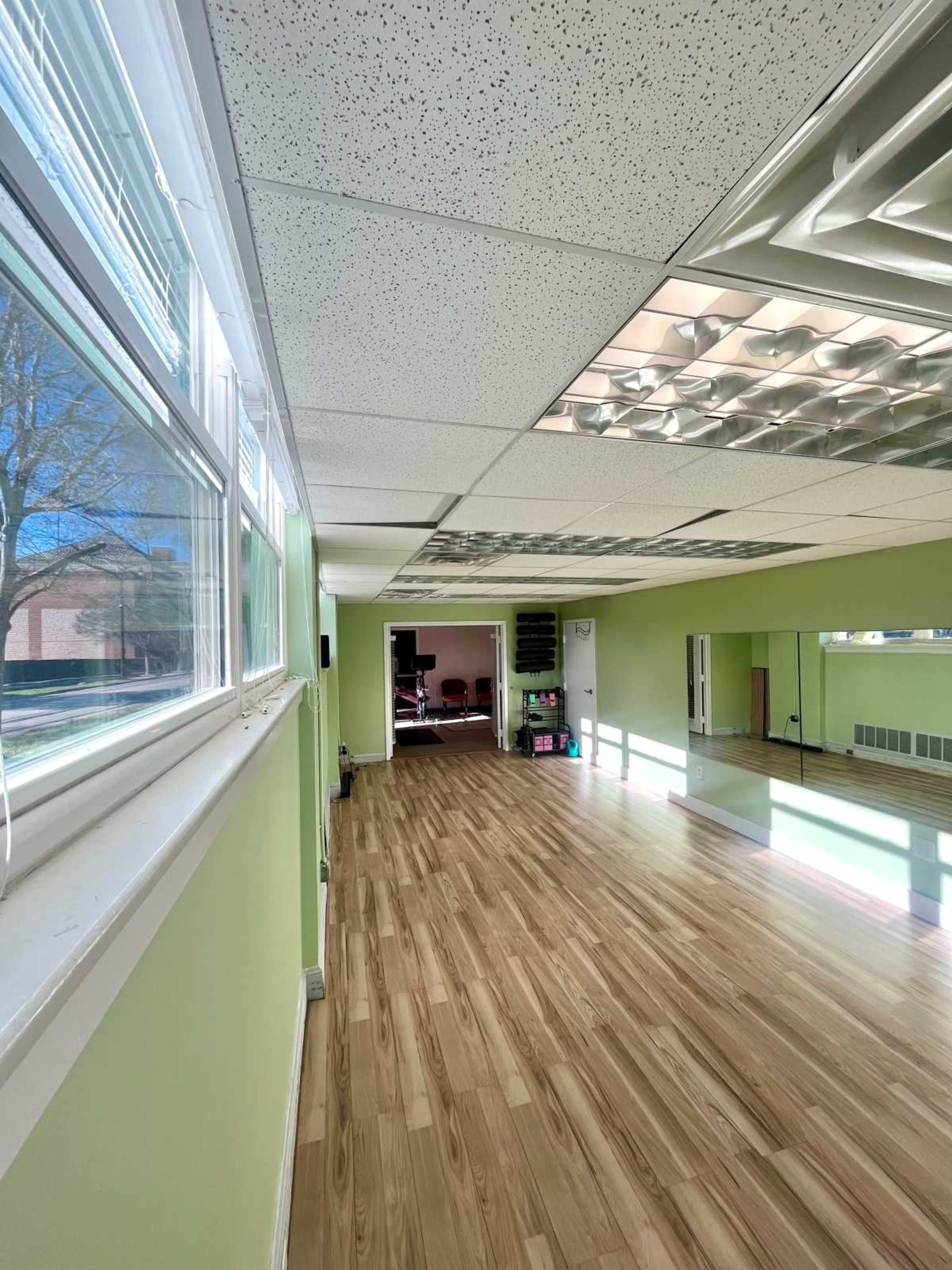 The image shows a spacious room with green walls, large windows, and a wooden floor, featuring mirrors along one side.