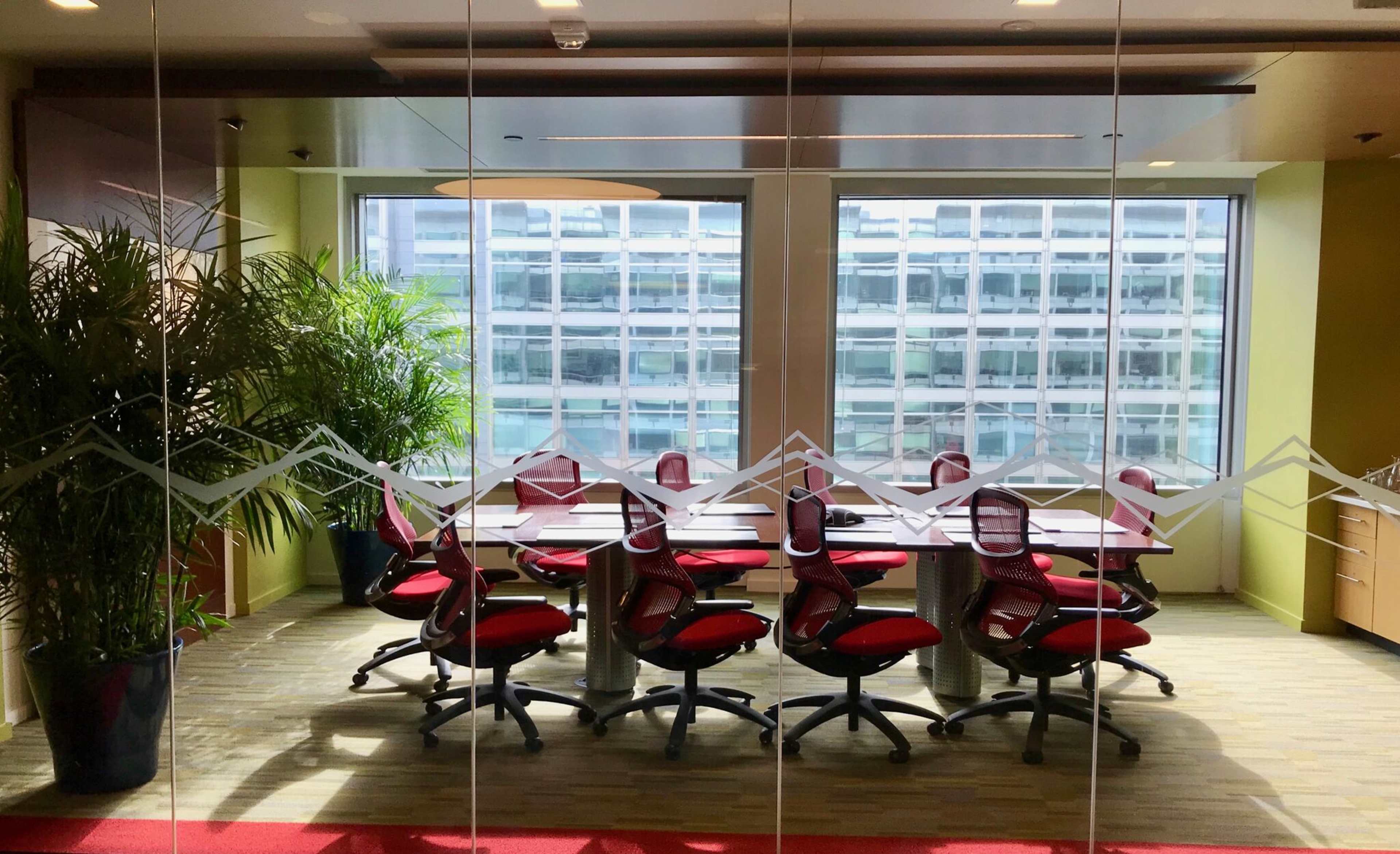A conference room with a long table surrounded by red office chairs and large windows overlooking a modern building.