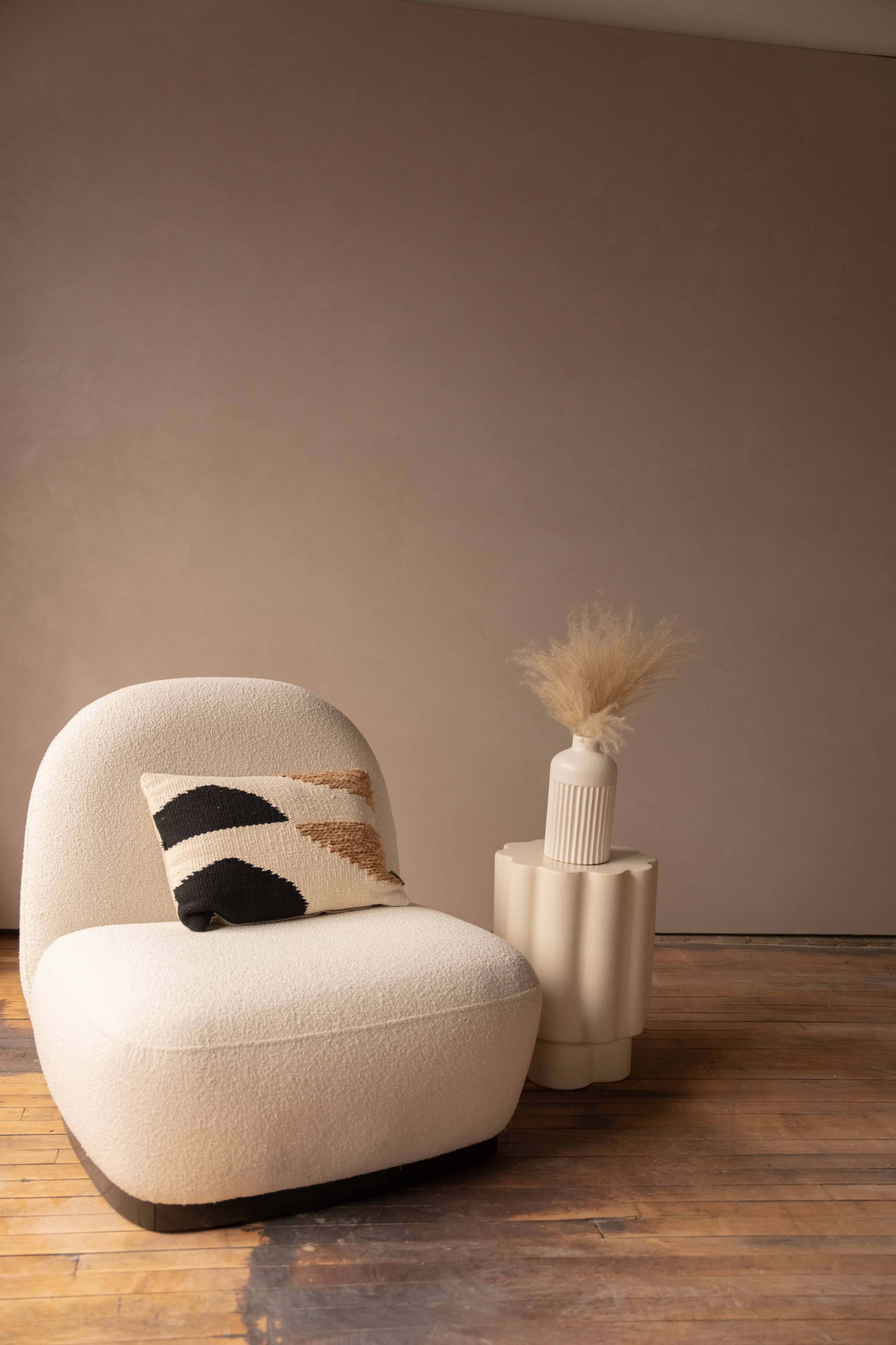 A curved, beige armchair with a patterned cushion sits next to a round, textured side table that holds a decorative vase with dried plants.