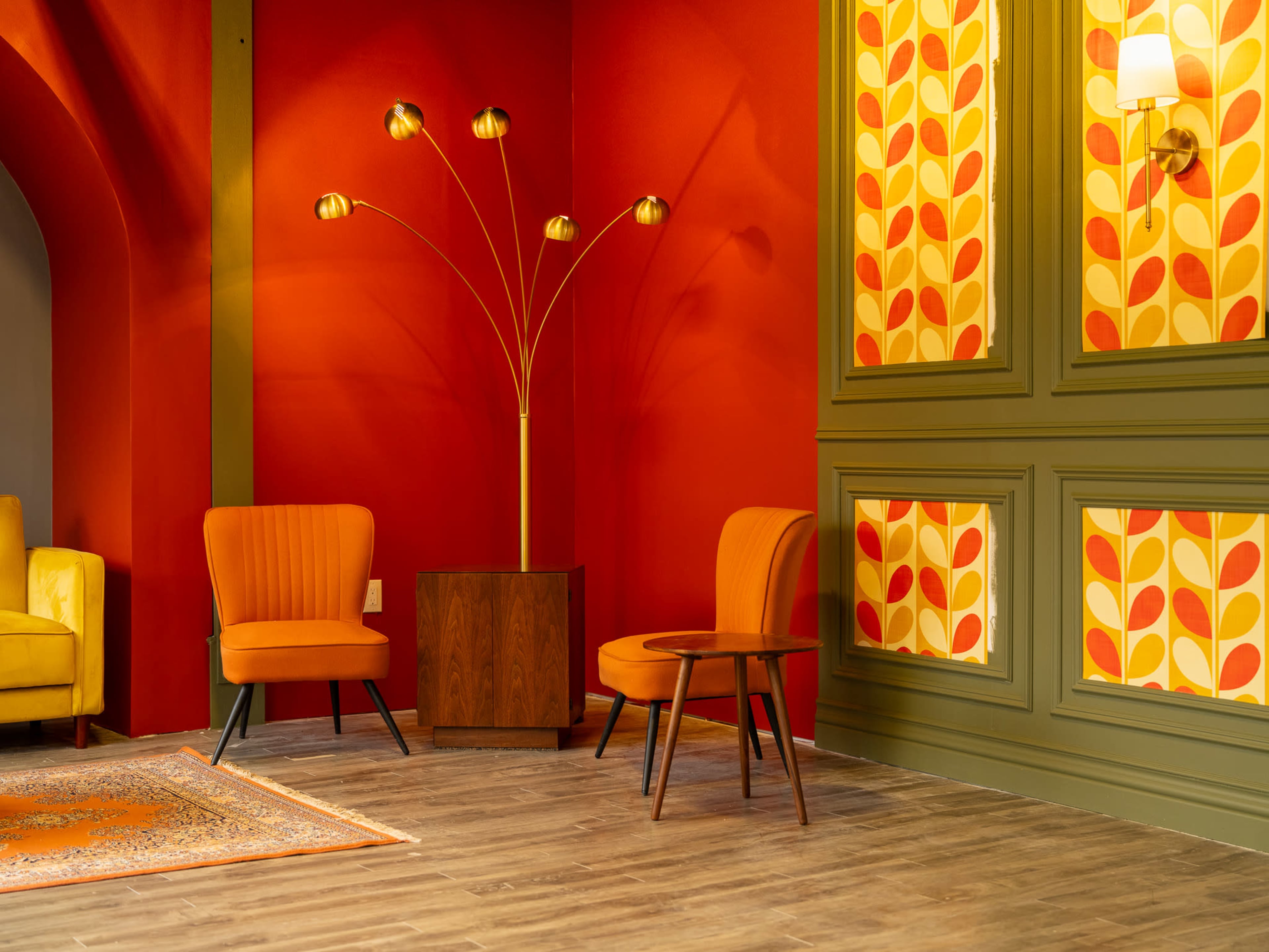 The image features a vibrant interior space with two orange armchairs, a wooden side table, and a decorative wall adorned with a retro floral pattern against a bold red backdrop.