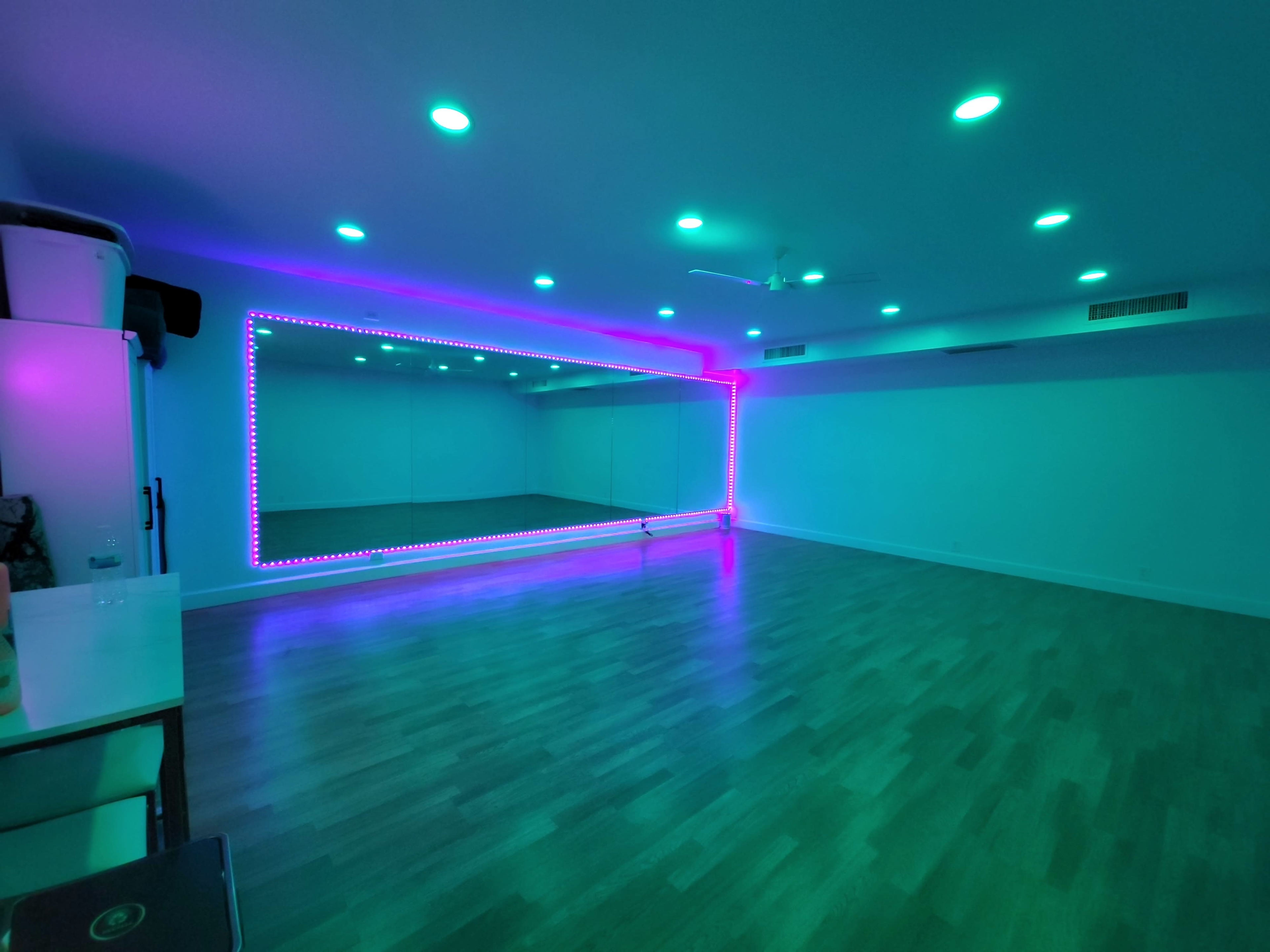 A spacious room with polished wood flooring and a mirrored wall framed by colorful LED lights.
