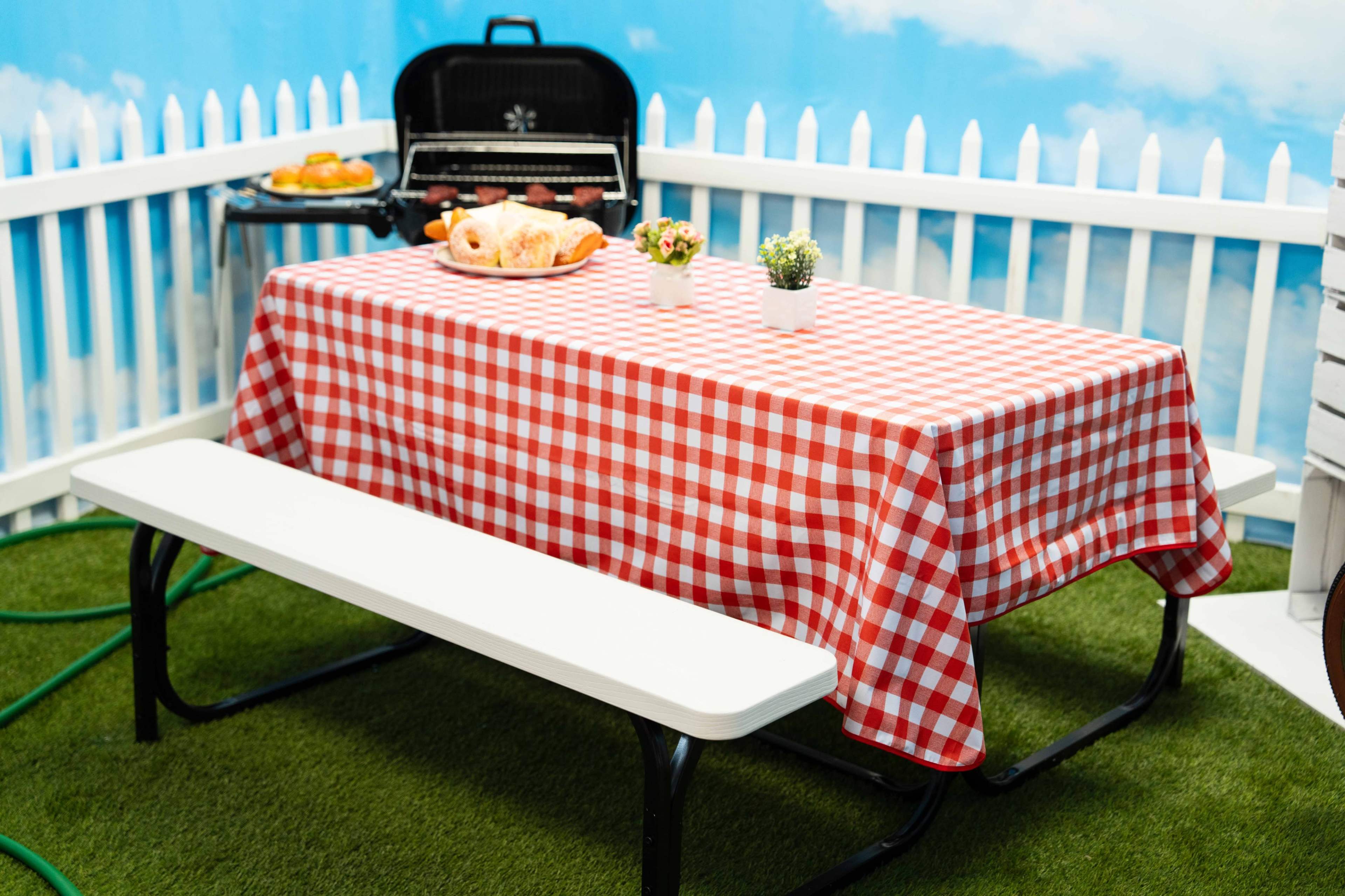 A picnic table covered with a red and white checked tablecloth is set up in a fenced area, accompanied by a grill and some food items.