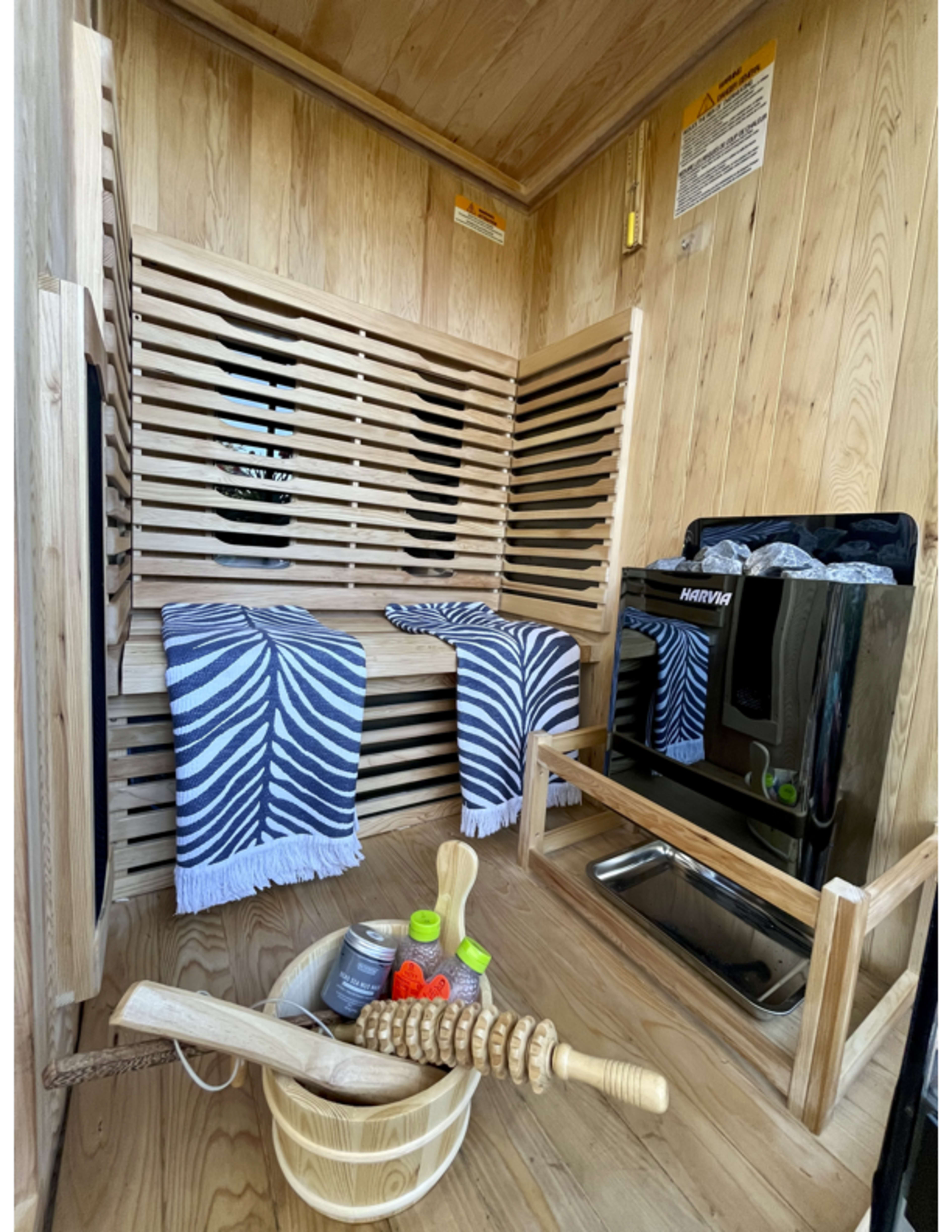 The image shows the interior of a wooden sauna featuring slatted seating, two blue and white towels, a black heater, a wooden bucket, a massage roller, and a few bottles.