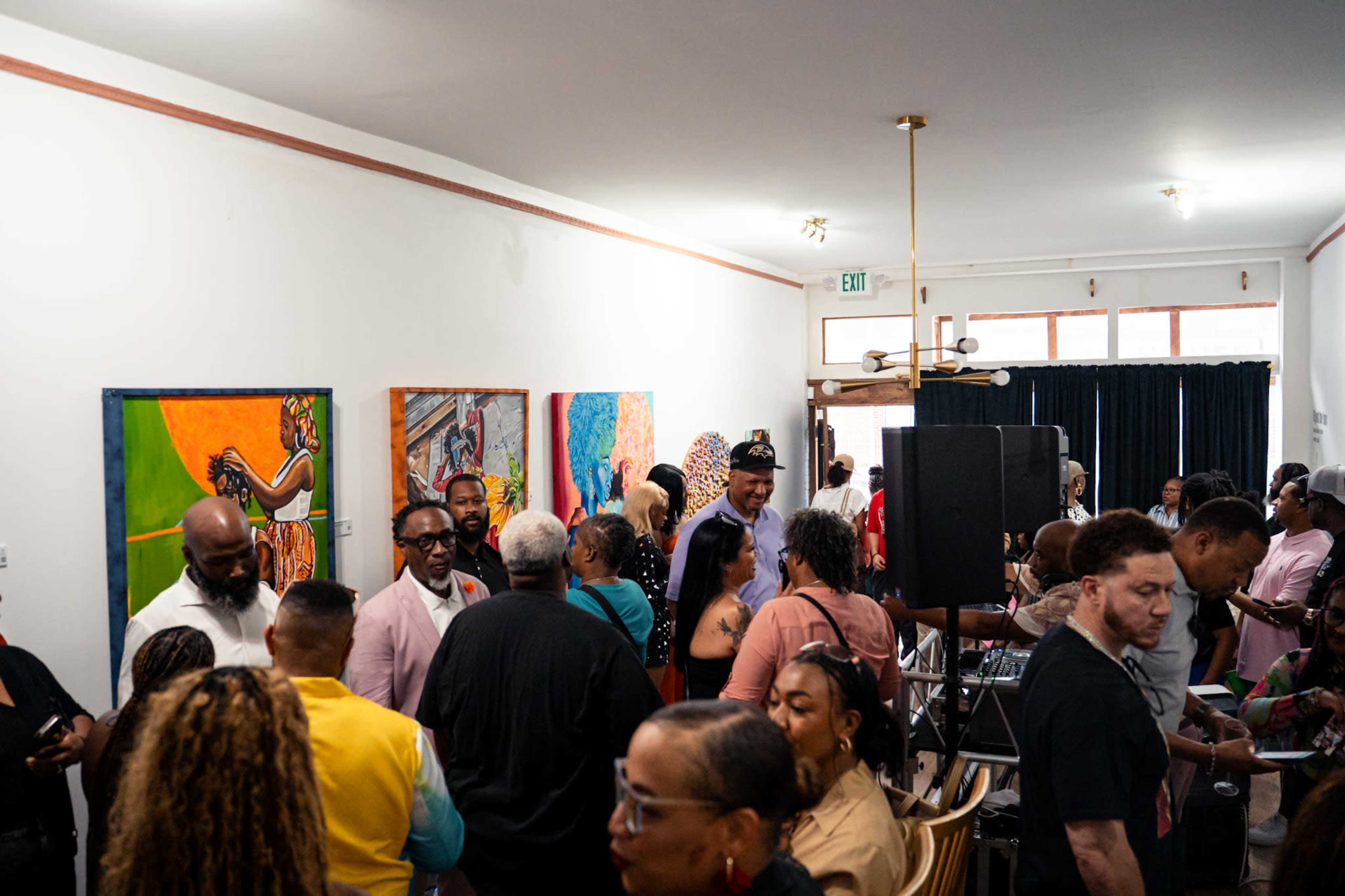 A crowded gallery space features people socializing and colorful artwork on the walls.