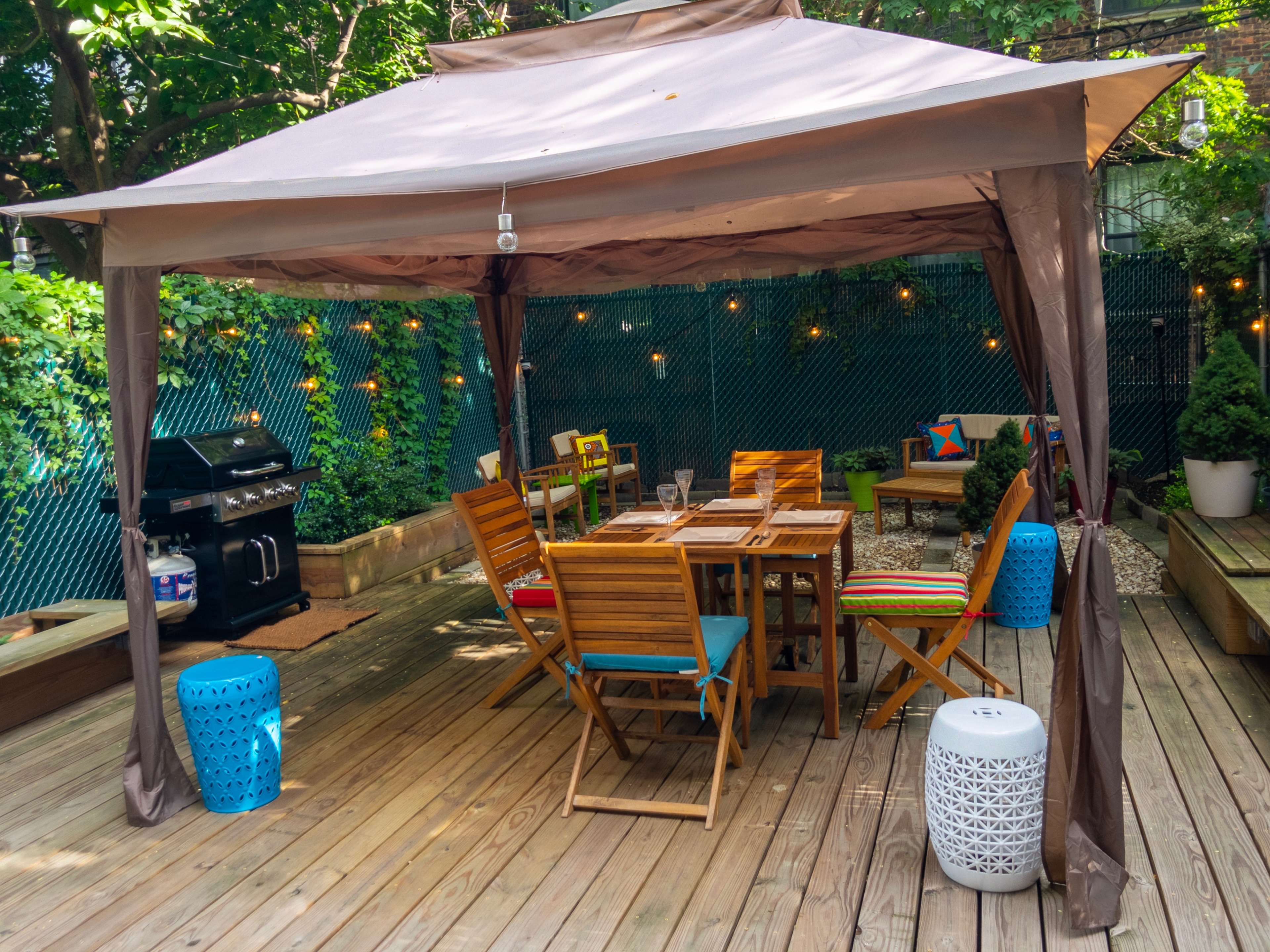 A wooden deck features a gazebo with a dining table, surrounded by colorful chairs, a barbecue grill, and decorative stools.