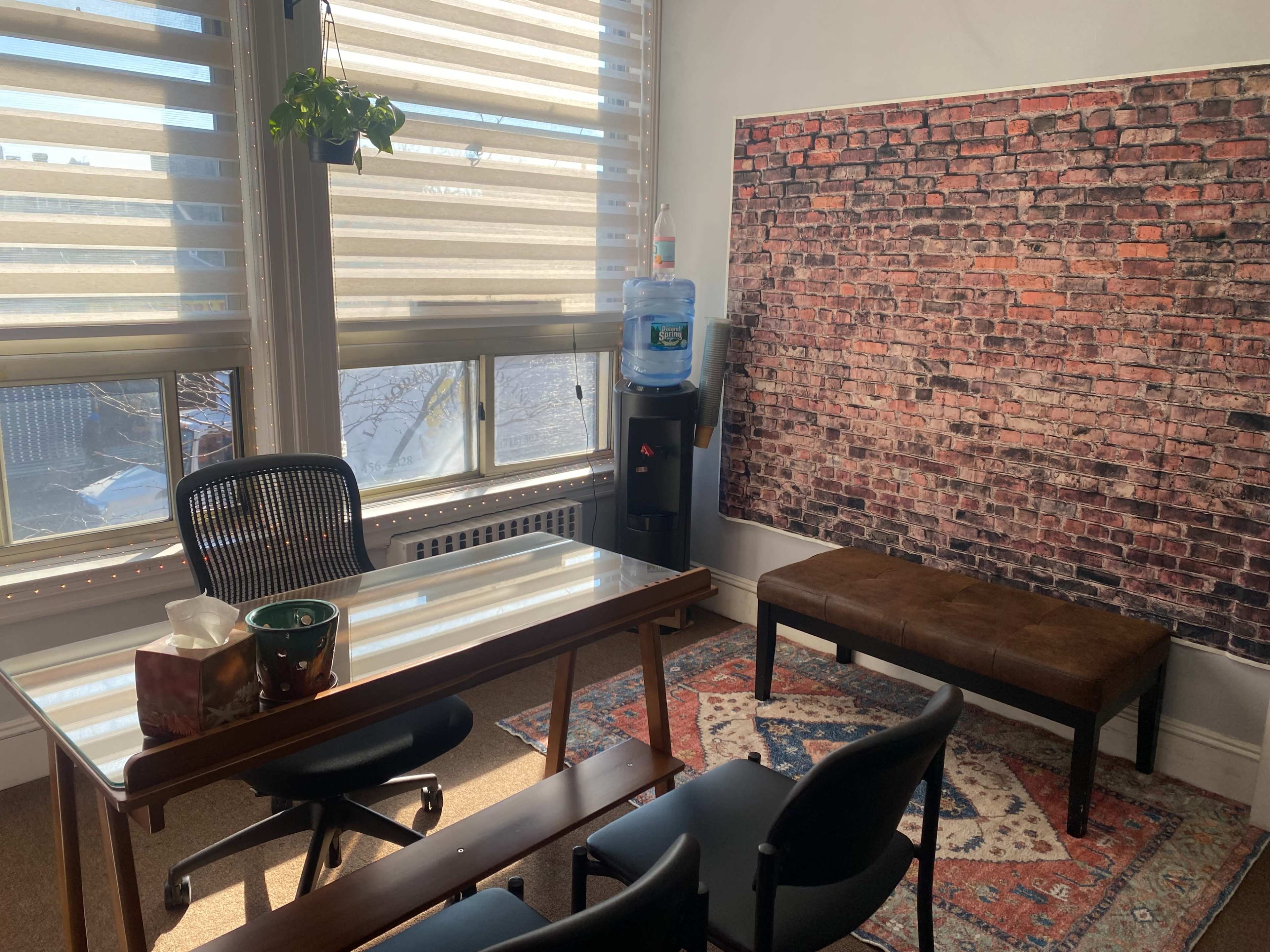 An office space featuring a glass desk, a plant in the window, a water cooler, and a brick-patterned wall behind a brown bench on a rug.