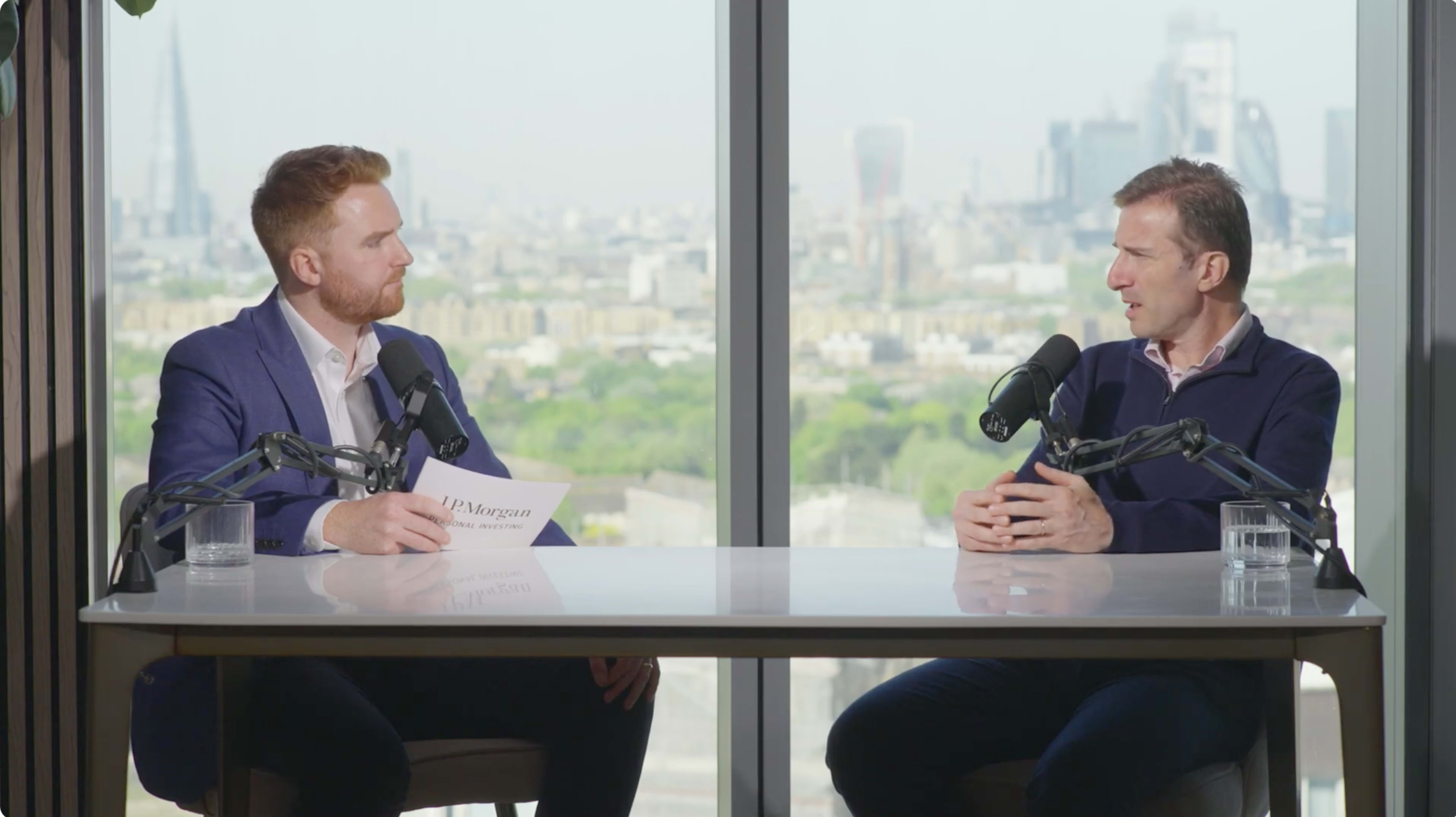 Two men are seated at a table with microphones, engaged in a conversation against a backdrop of a city skyline.