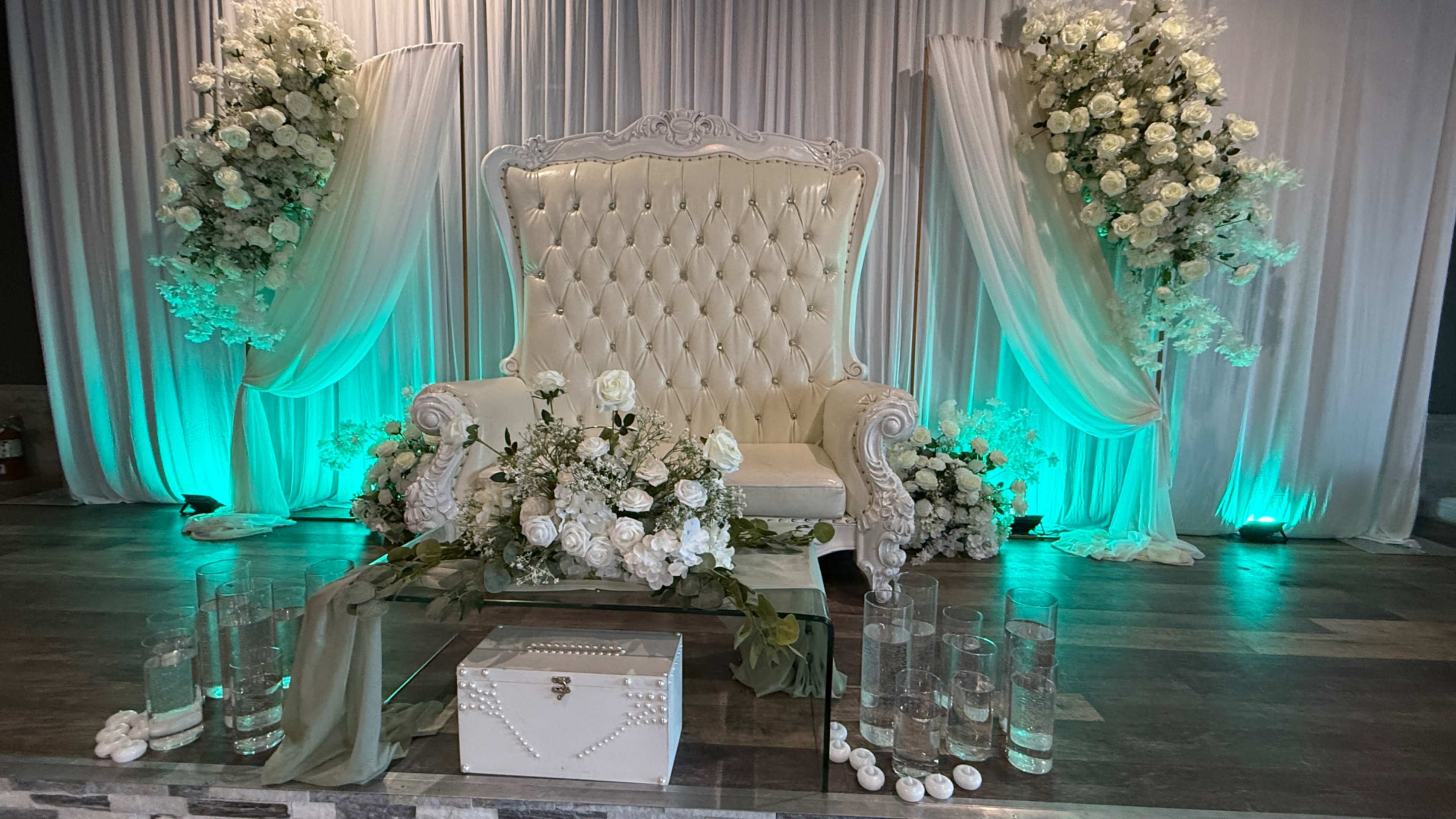 The image shows an elegantly decorated event space featuring a large white throne chair flanked by floral arrangements and lit with green ambient lighting.