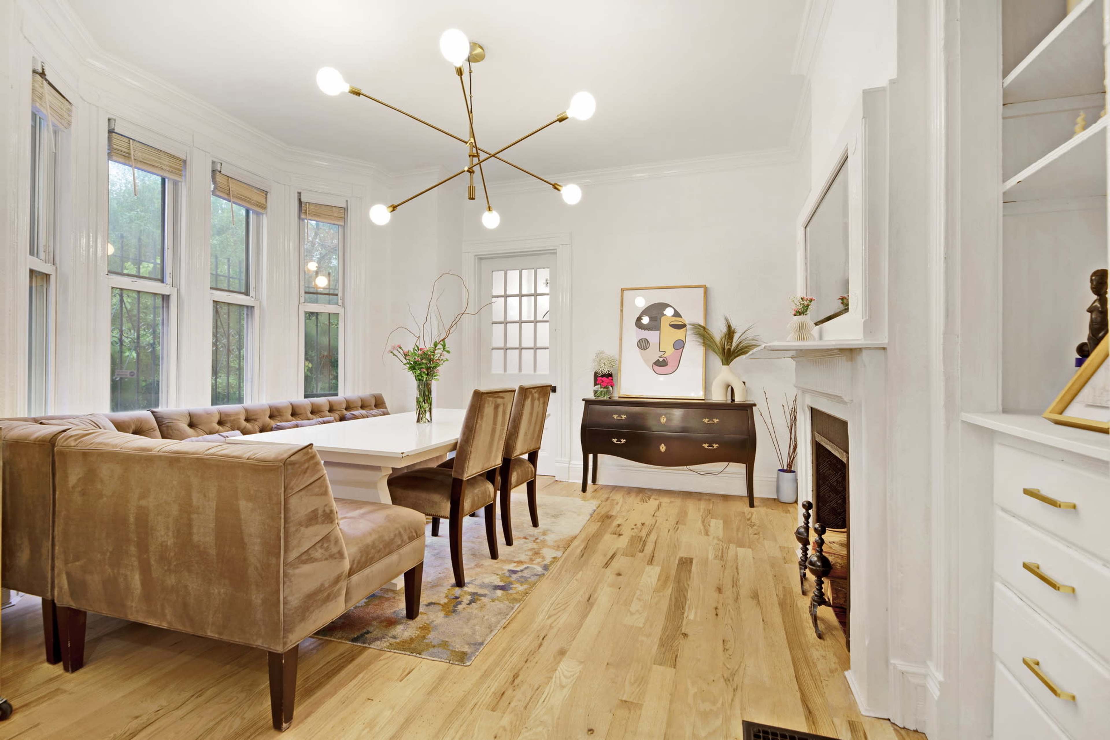 A dining room features a long white table surrounded by six brown velvet chairs, with large windows allowing natural light and a modern chandelier overhead.