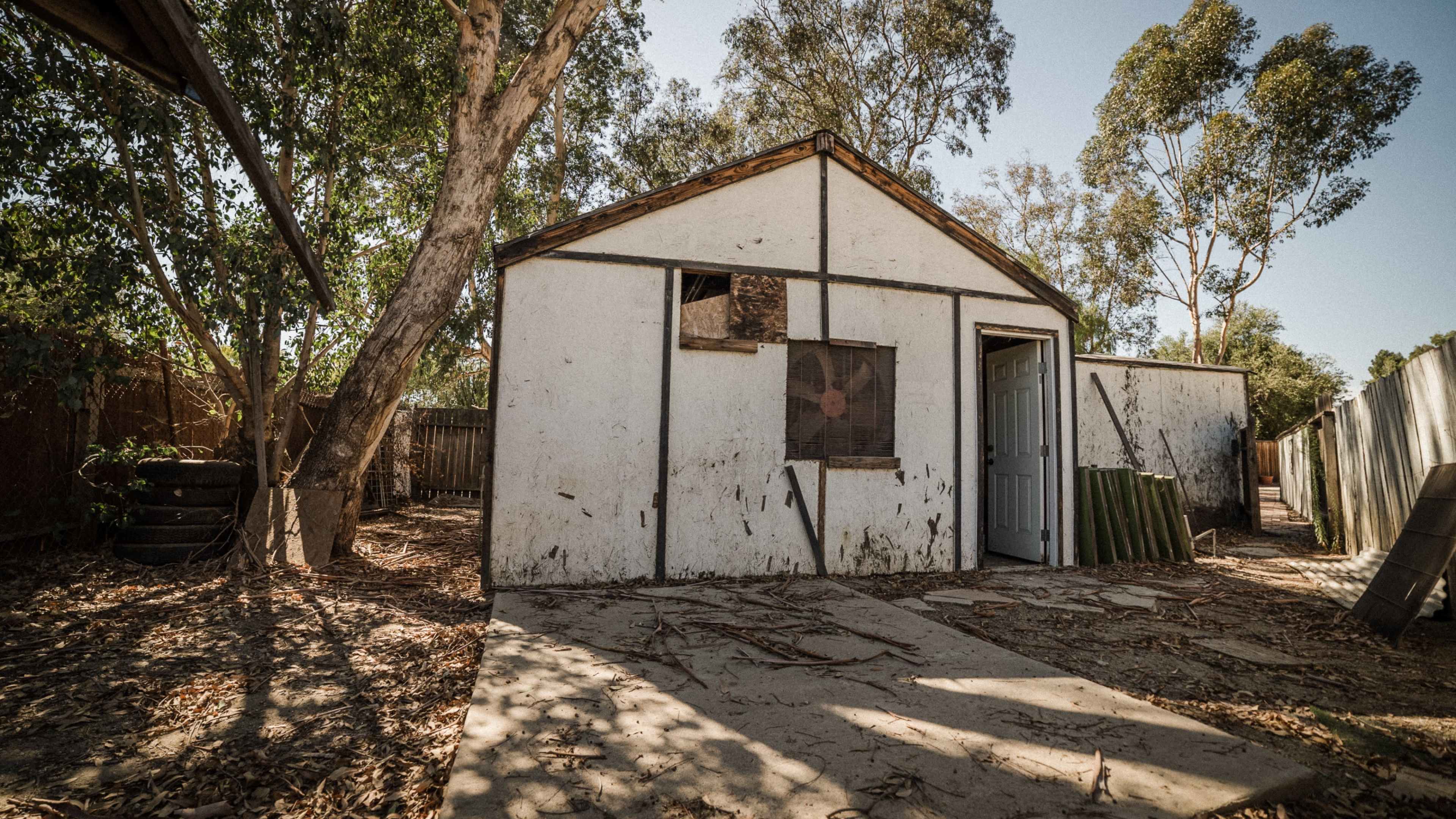 A weathered, white shed with a door and a window, surrounded by dirt and sparse vegetation.