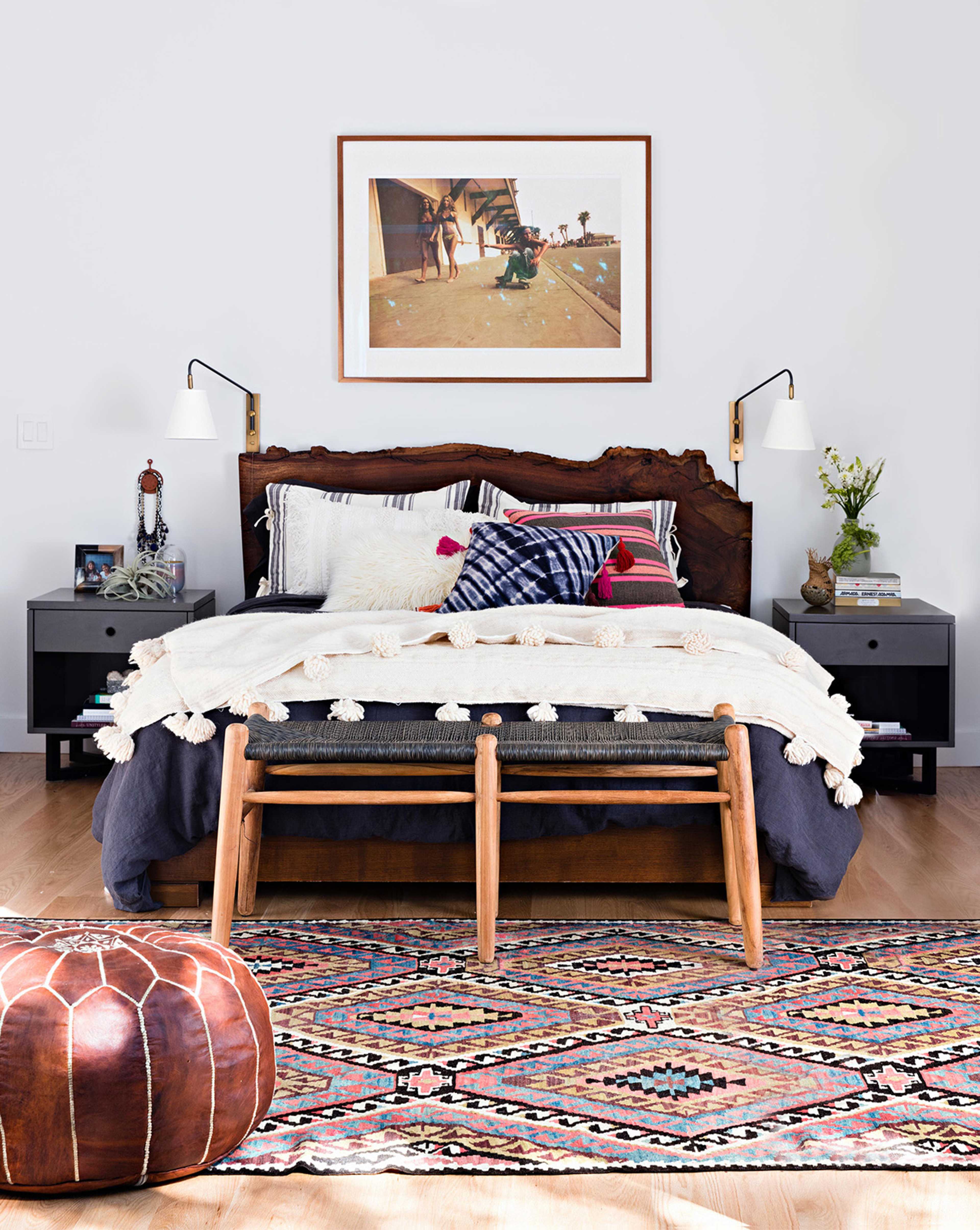 A cozy bedroom features a wooden bed with decorative pillows, a knitted blanket, and a patterned rug, accompanied by bedside tables and wall-mounted lamps.