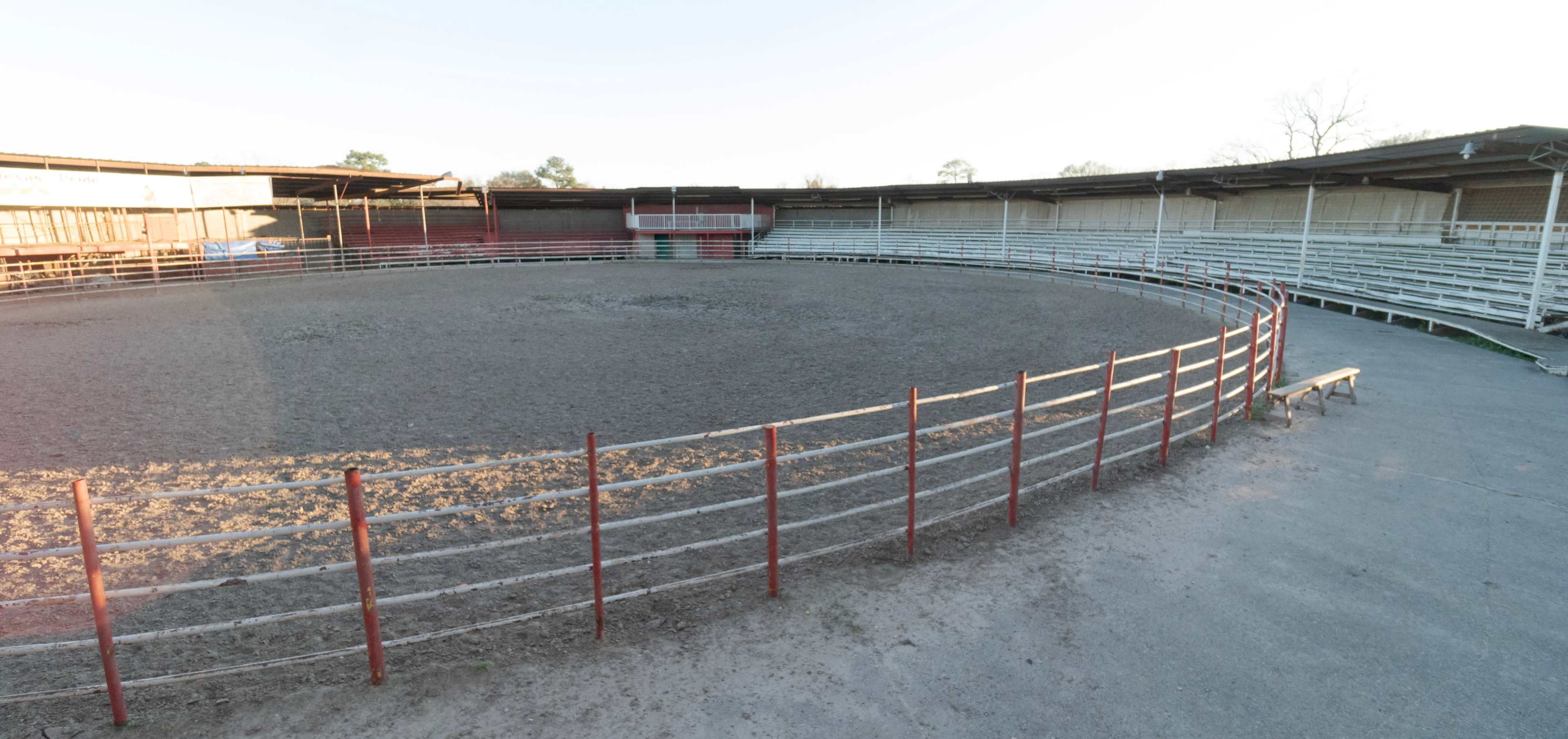 An empty rodeo arena with a circular dirt floor surrounded by a white wooden fence and bleacher seating.