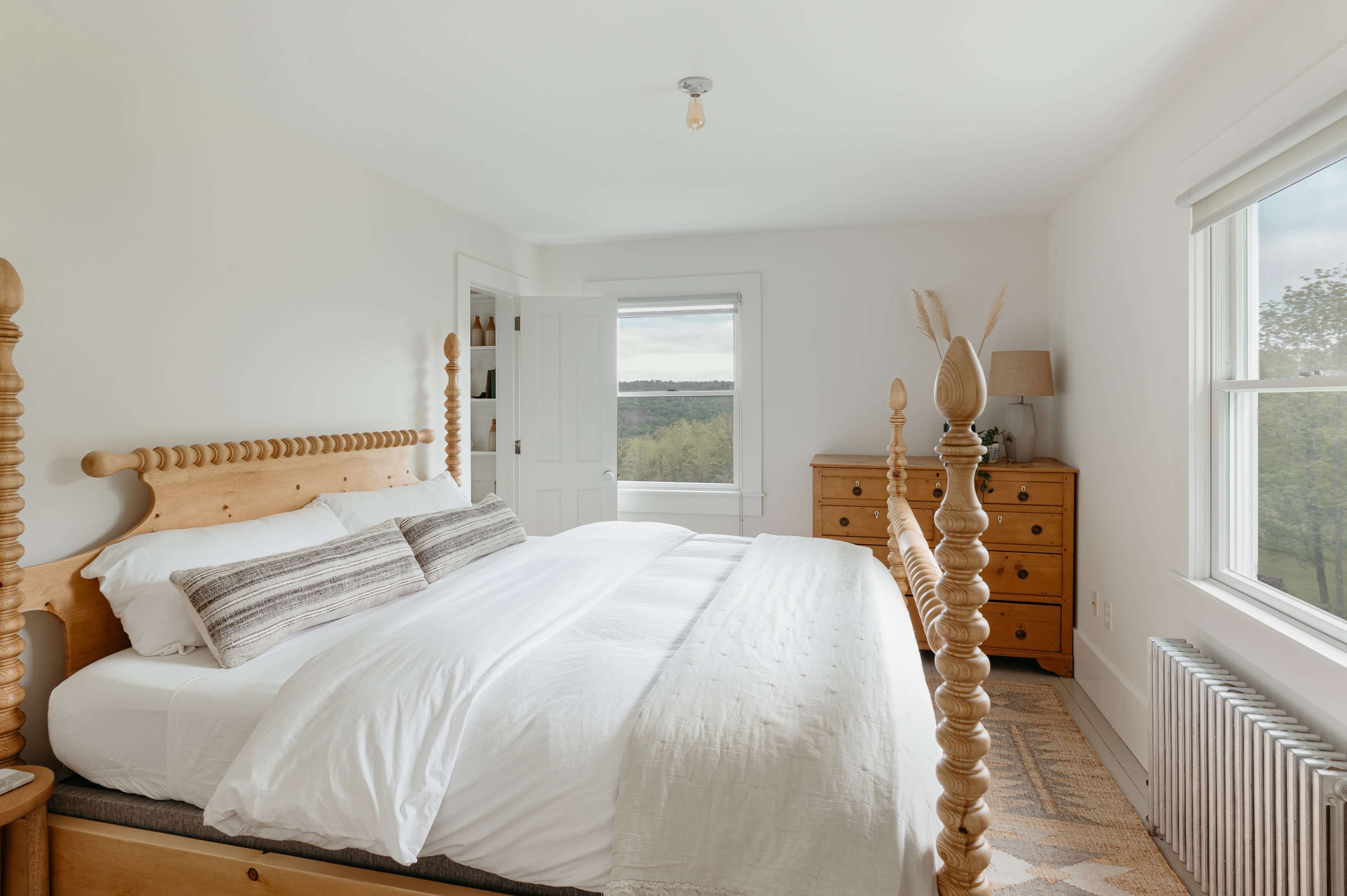 A bright, minimalist bedroom featuring a wooden four-poster bed, white bedding, a dresser, and a large window with a view of greenery.
