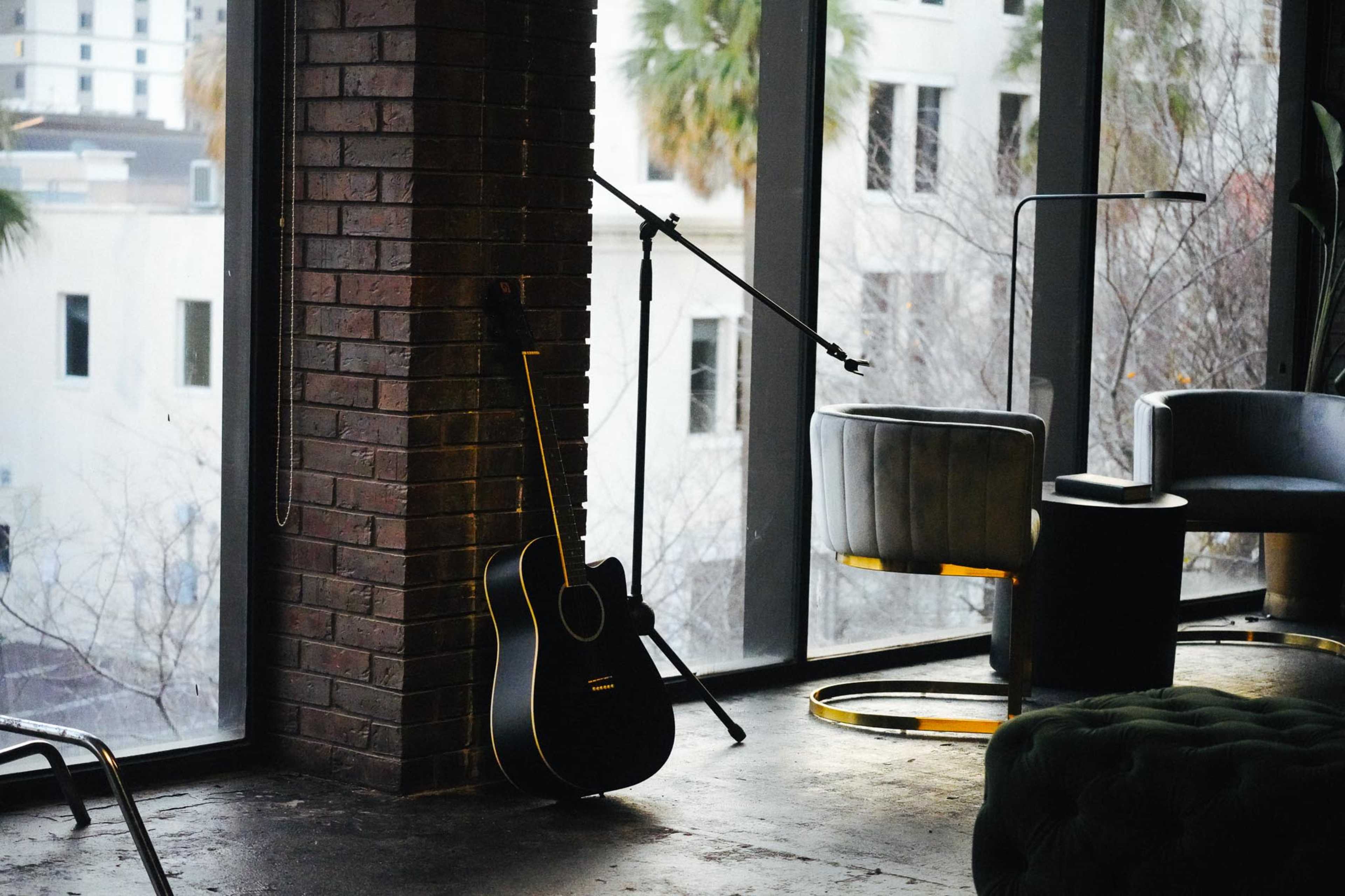A black acoustic guitar leans against a brick wall near a large window, beside a microphone stand and modern seating.