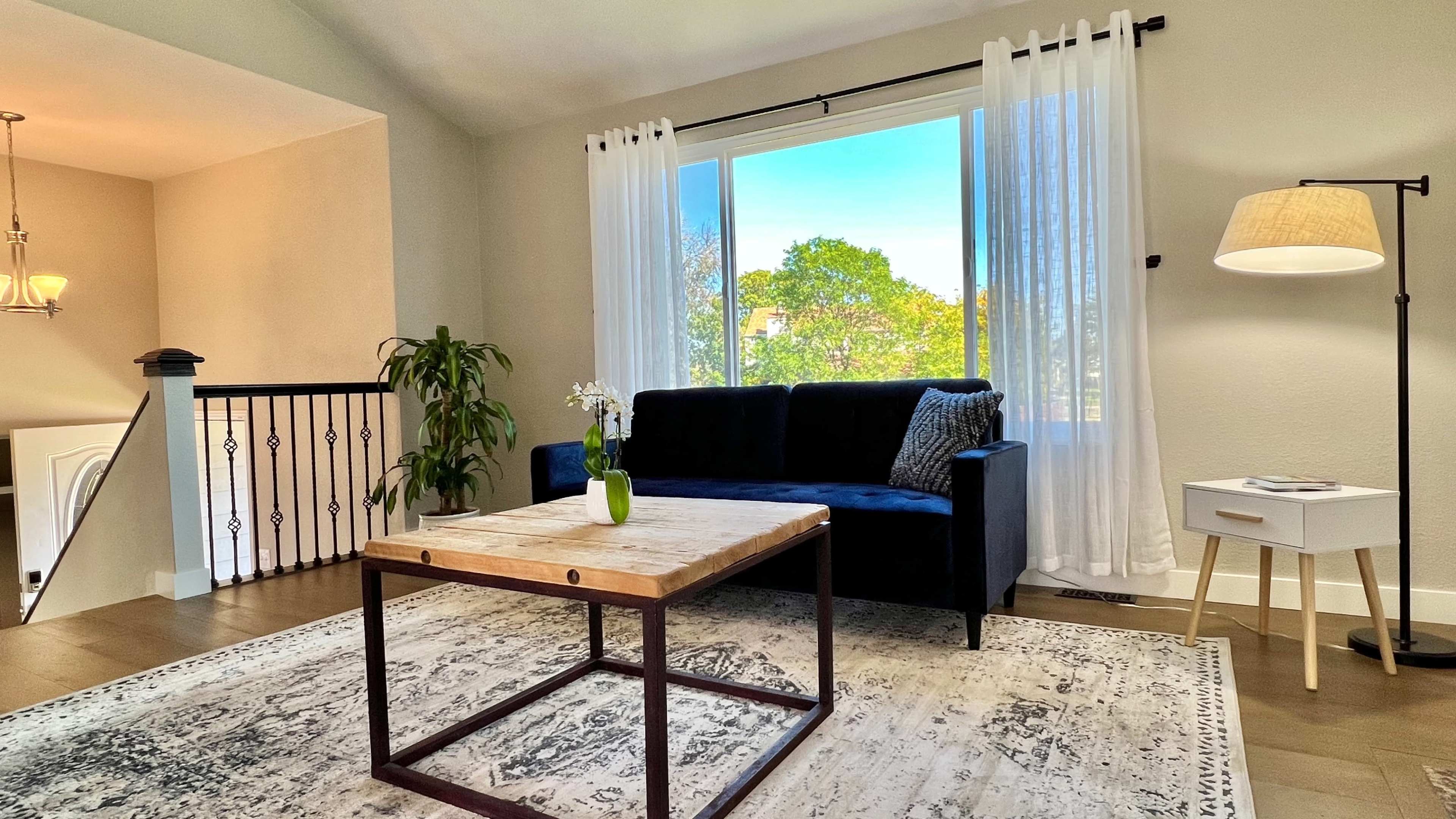 The room features a blue sofa, a wooden coffee table, and large windows with sheer curtains, allowing natural light to fill the space.