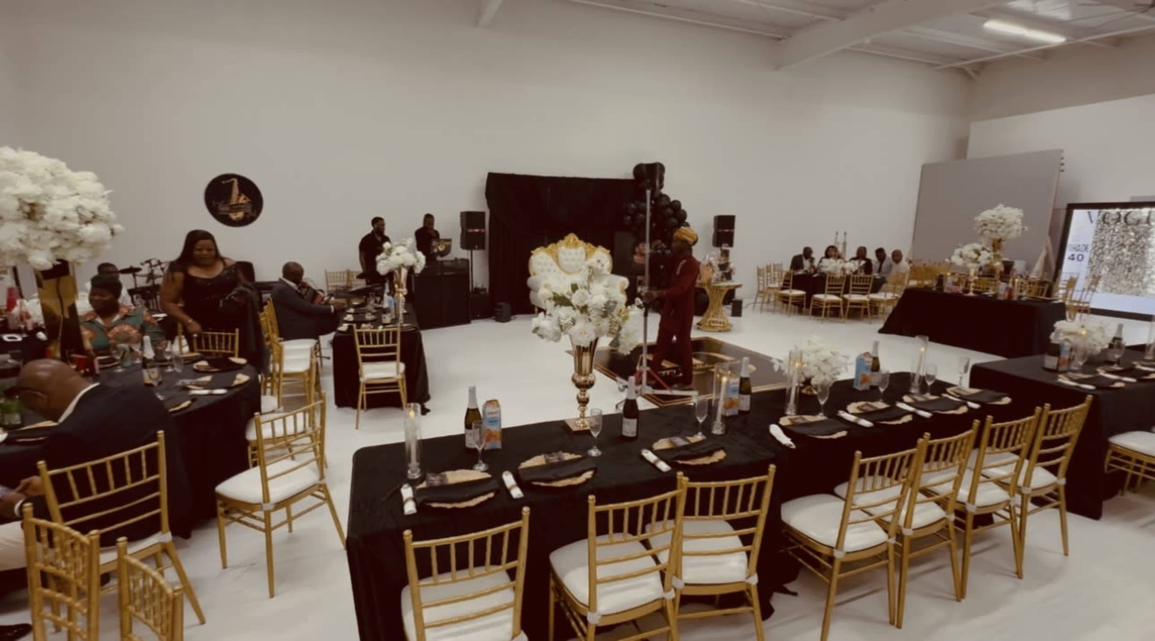 The image shows a banquet hall set for an event, featuring tables adorned with black tablecloths and gold chairs, decorated with white floral arrangements.